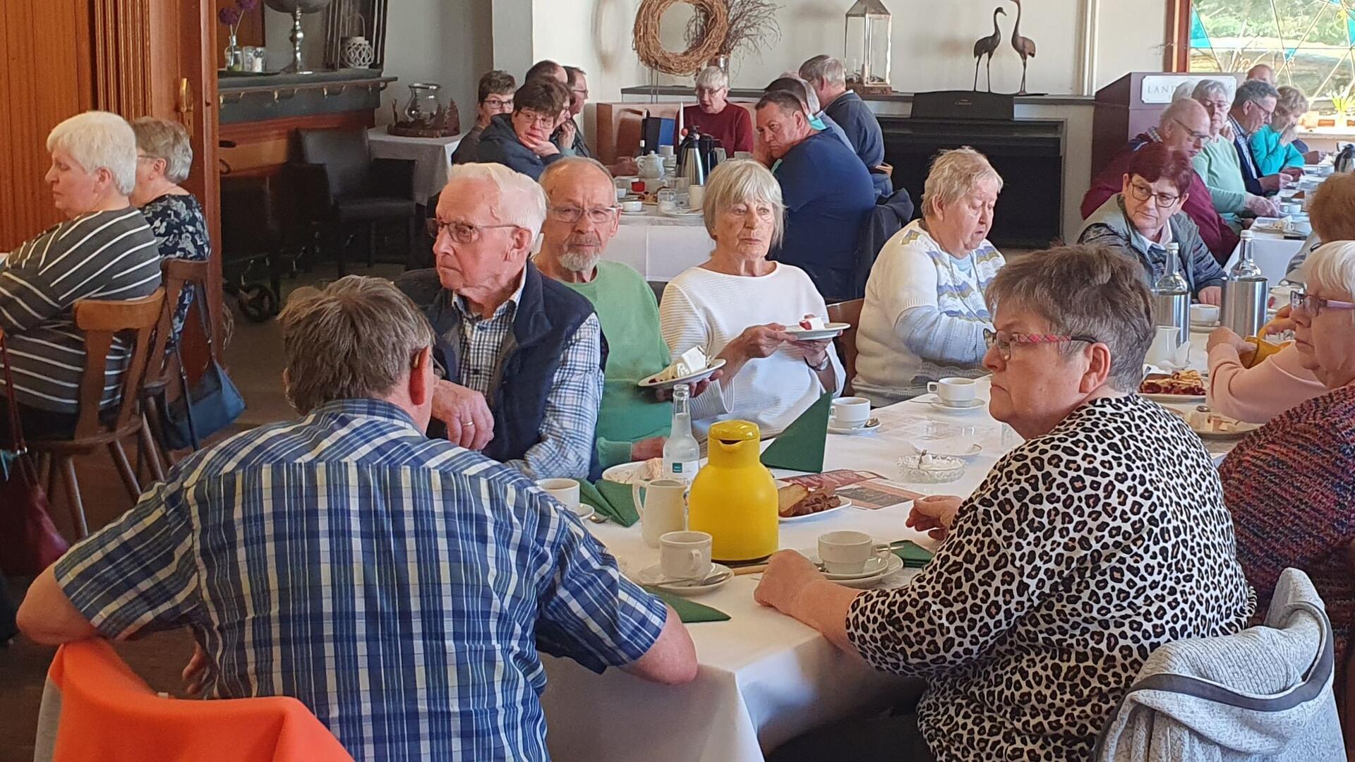 Überwiegend ältere Menschen sitzen an gedeckten Tischen, genießen Kaffee und Kuchen und scheinen einer Ansprache zu lauschen. 