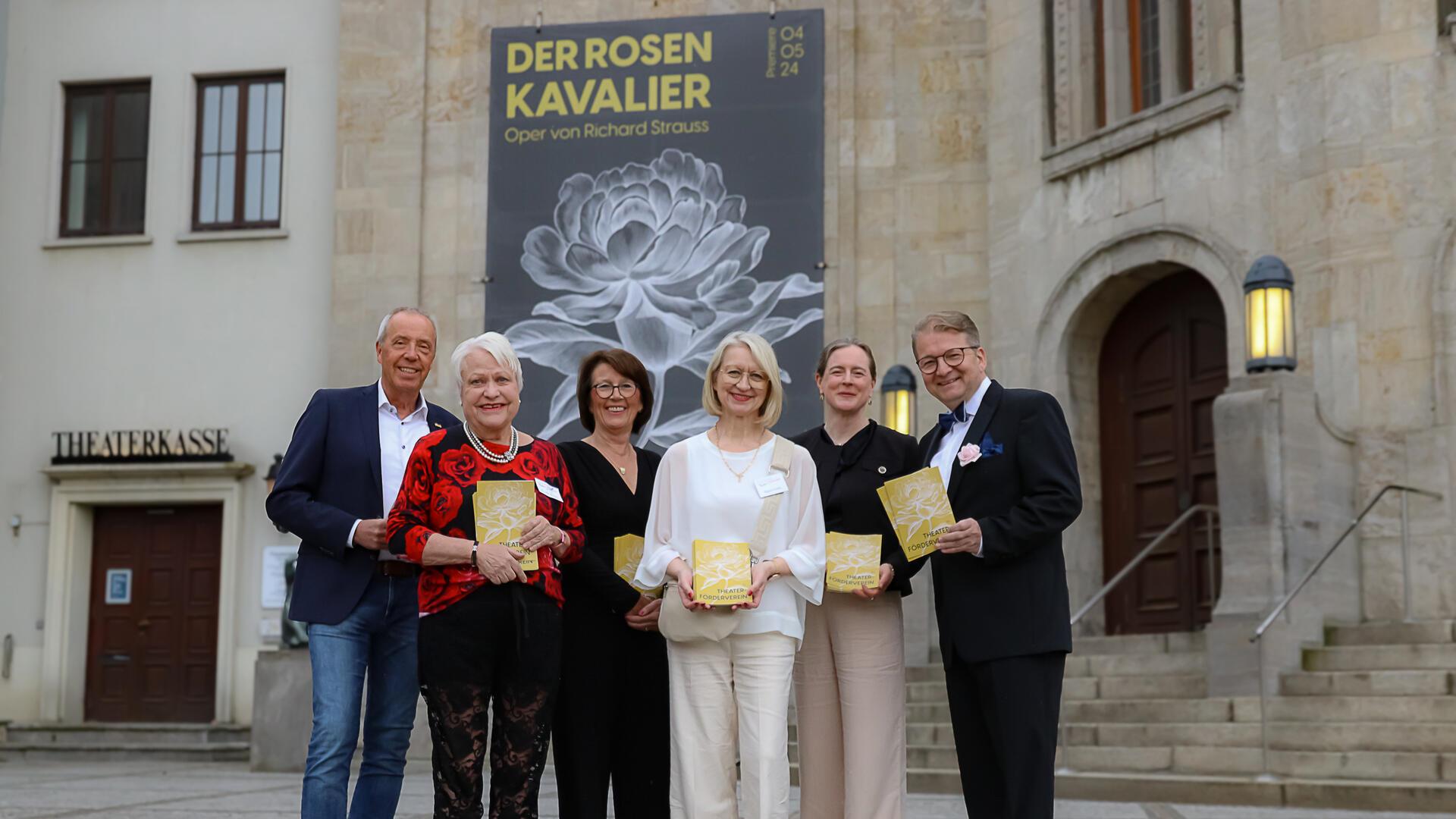 vier Frauen und zwei Männer stehen vor dem Eingang des Stadttheaters Bremerhaven, an dessen Fassade ein großes Plakat hängt mit der Aufschrift "Der Rosenkavalier – Oper von Richard Strauss"