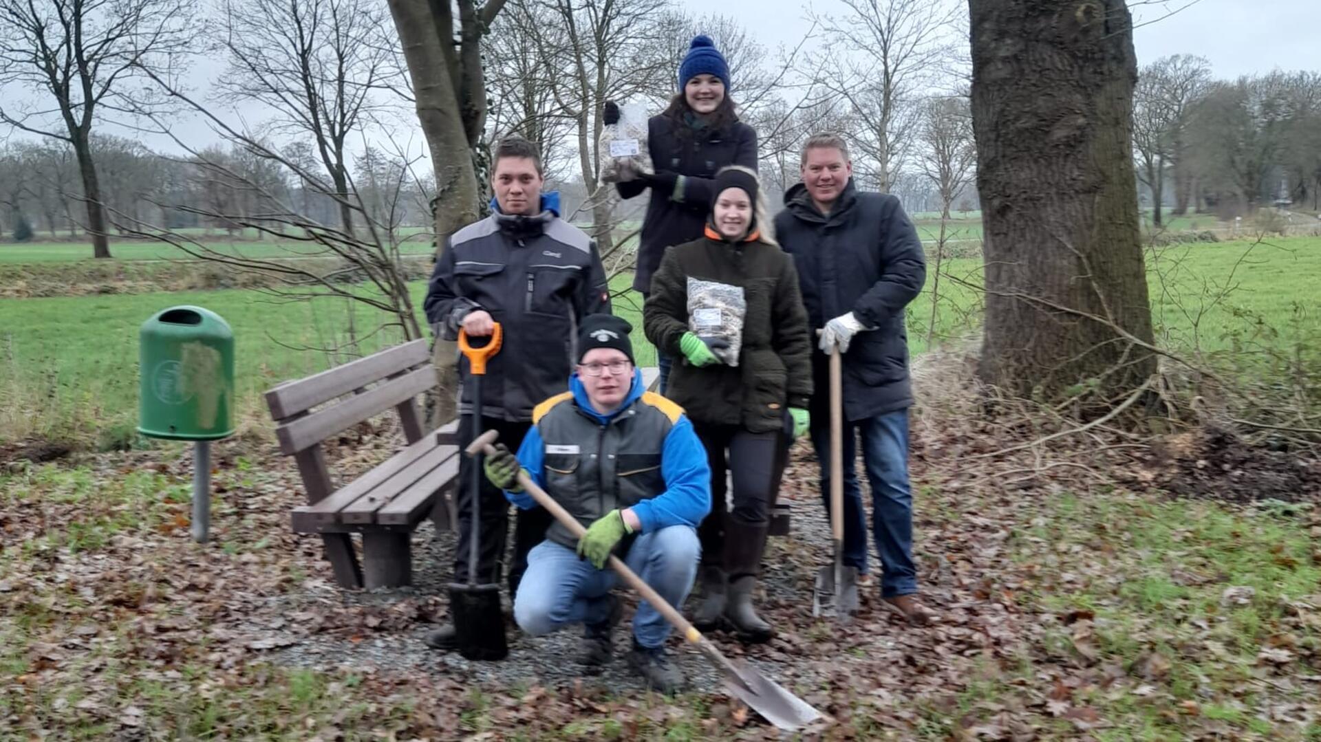 Eine Gruppe von fünf Personen steht und kniet in einem ländlichen Gebiet vor einer Sitzbank. Sie tragen warme Kleidung und Arbeitshandschuhe, passend für Garten- oder Landschaftsarbeiten. Einige halten Schaufeln und Gartengeräte in der Hand.