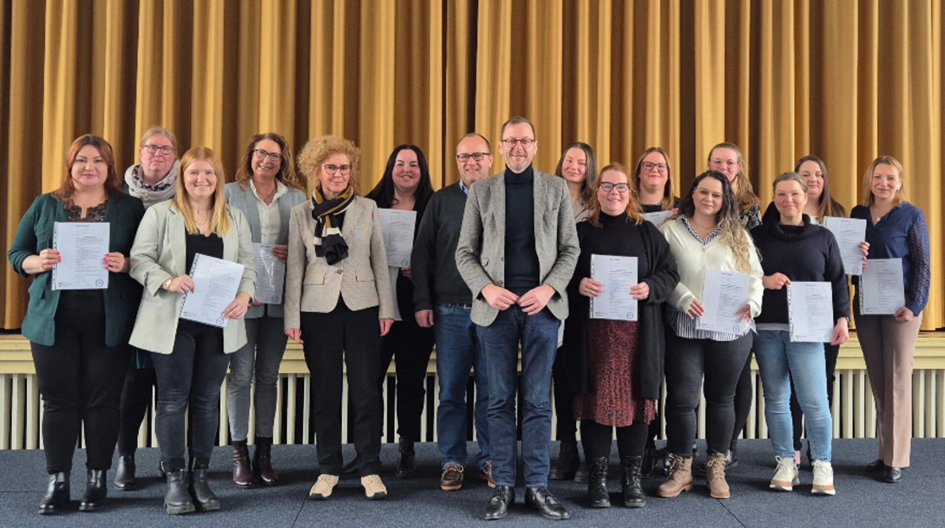 Eine Gruppe von 17 Personen, überwiegend Frauen, posiert auf einer Bühne vor einem goldfarbenen Vorhang. Viele der Personen halten Zertifikate oder Dokumente in der Hand und lächeln in die Kamera. Die Gruppe ist in gepflegter, teils geschäftlicher Kleidung gekleidet. 