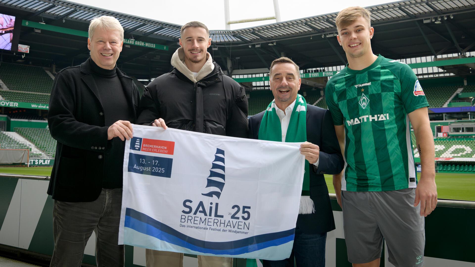 Das Foto ist im Weserstadion entstanden, es zeigt vier Herren, die eine Flagge in der Hand halten, die auf die Sail 2025 hinweist. Freuen sich über die Partnerschaft: Dr. Ralf Meyer vom SAiL-Organisationskomitee, die Spieler Marco Friedl und Keke Topp sowie Michael Gerber, Geschäftsführer der Erlebnis Bremerhaven GmbH.