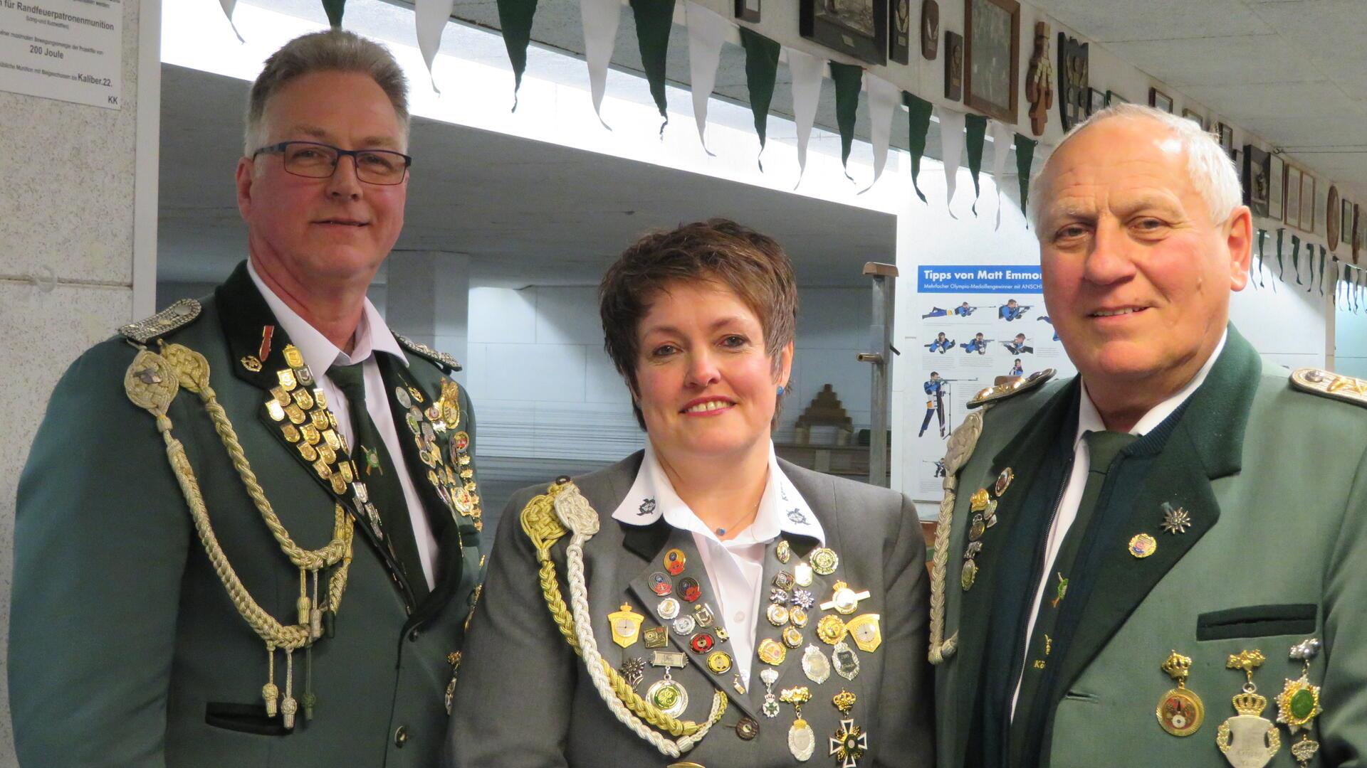Drei Mitglieder eines Schützenvereins in festlicher Uniform mit zahlreichen Orden und Abzeichen stehen nebeneinander und lächeln in die Kamera. Im Hintergrund sind grün-weiße Wimpelketten und Schießstand-Zubehör zu sehen.