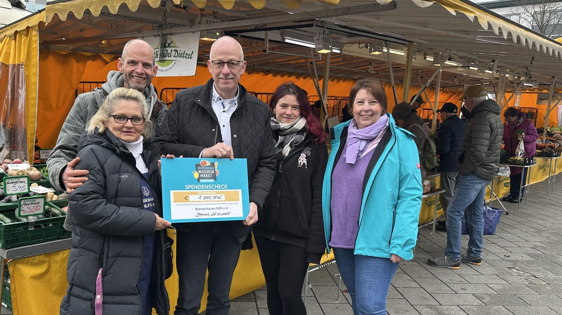 Fünf Personen stehen auf einem Wochenmarkt vor einem Gemüsestand und halten gemeinsam einen großen Spendenscheck über 1.000 Euro in die Kamera. Im Hintergrund sind Marktstände mit gelben Planen und weitere Besucherinnen und Besucher zu sehen.
