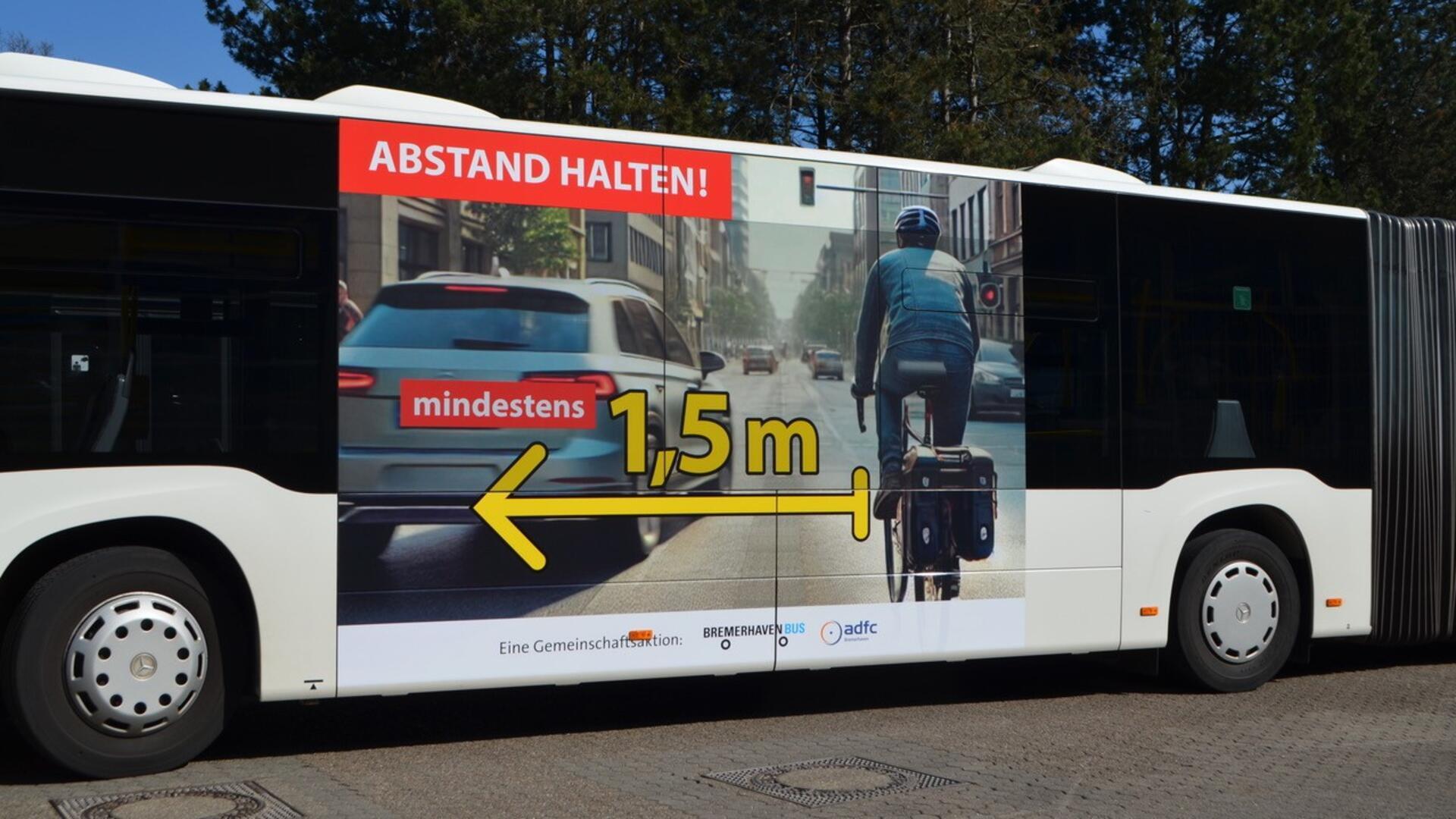 Ein Linienbus zeigt auf seiner Seitenfläche eine große Werbegrafik mit dem Hinweis „ABSTAND HALTEN!“. Die Abbildung zeigt ein Auto neben einem Radfahrer in der Stadt. Dazwischen ist ein gelber Doppelpfeil mit der Aufschrift „mindestens 1,5 m“ zu sehen.