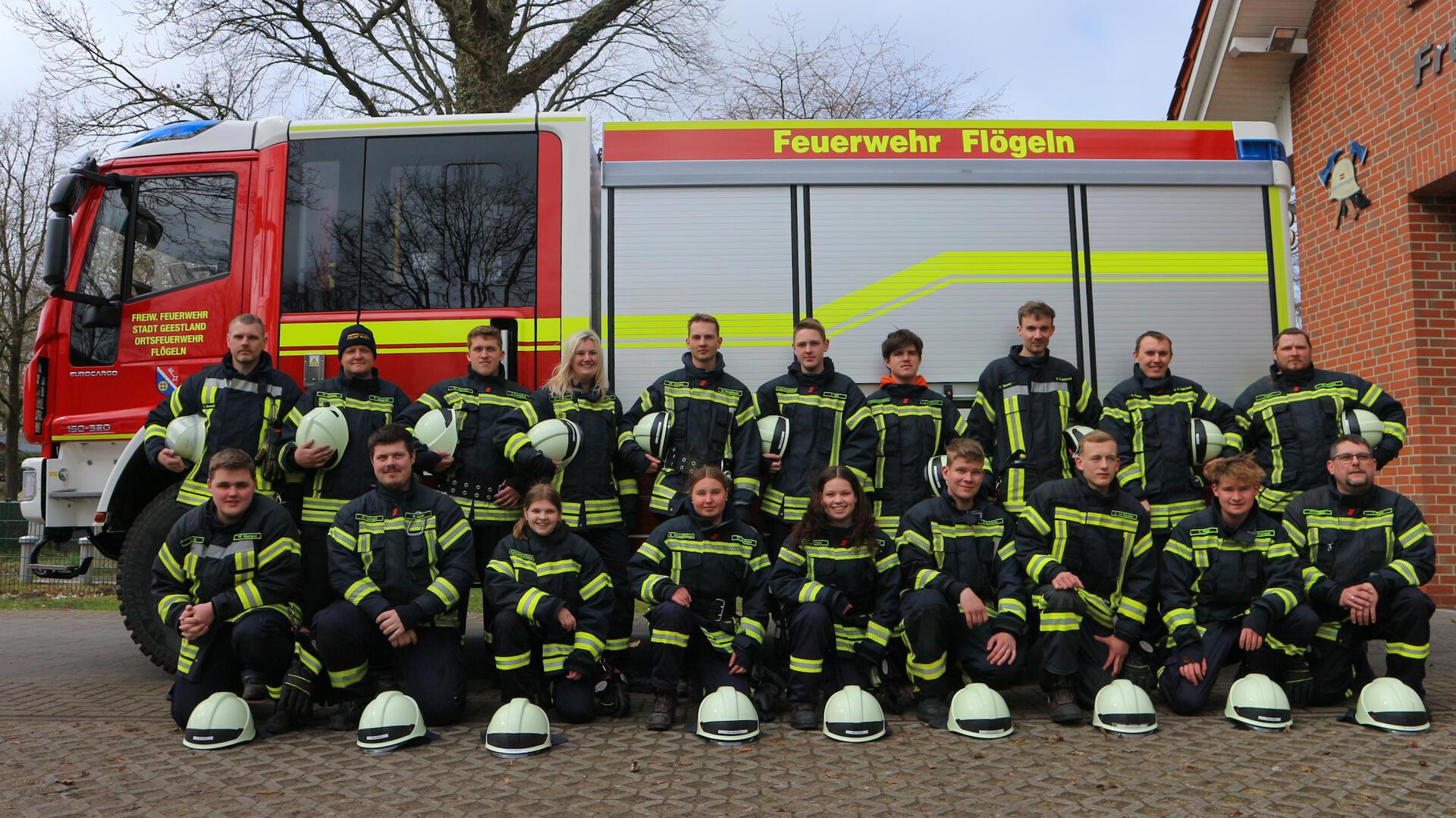 Gruppenfoto der Freiwilligen Feuerwehr vor einem Einsatzfahrzeug der Feuerwehr Flögeln. Die Feuerwehrleute tragen vollständige Schutzkleidung, viele halten ihre Helme in den Händen oder haben sie vor sich auf den Boden gestellt. Im Hintergrund sind das Feuerwehrfahrzeug und ein Teil des Feuerwehrhauses zu sehen.