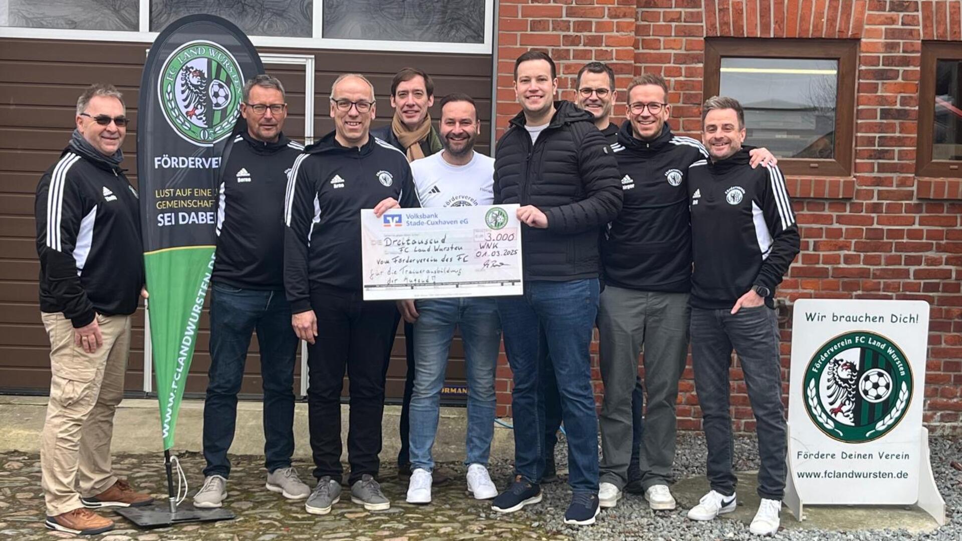 Eine Gruppe von Männern steht lächelnd vor einem Gebäude aus rotem Klinkerstein. Sie halten gemeinsam einen großen symbolischen Spendenscheck über 3.000 Euro, ausgestellt vom Förderverein des FC Land Wursten. Mehrere Männer tragen Vereinskleidung mit Vereinslogo. 