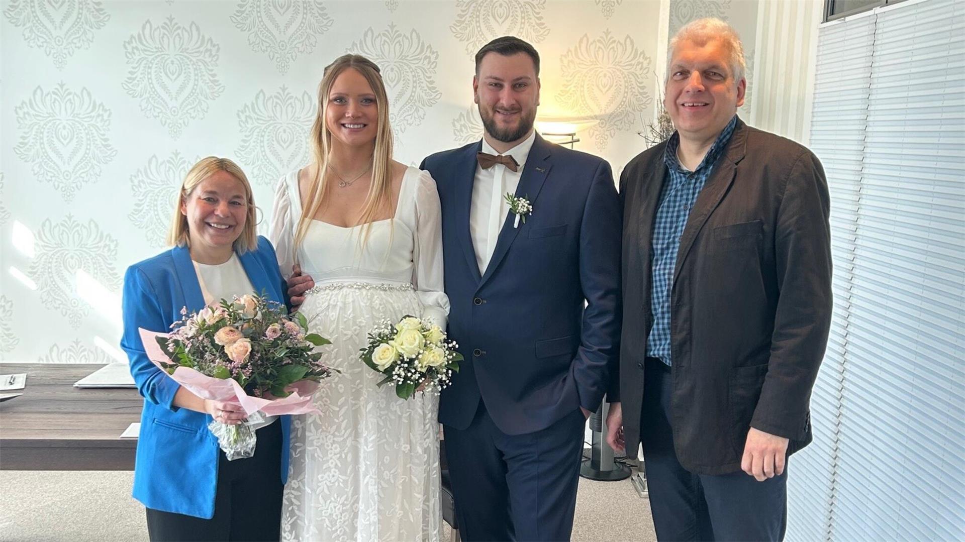 Vier Personen posieren fröhlich für ein Foto bei einer standesamtlichen Hochzeit. In der Mitte stehen das Brautpaar – die Braut in weißem Kleid mit Blumenstrauß, der Bräutigam im blauen Anzug mit Fliege. Links steht eine Frau mit blauem Blazer und einem weiteren Blumenstrauß, rechts ein Mann im dunklen Sakko.