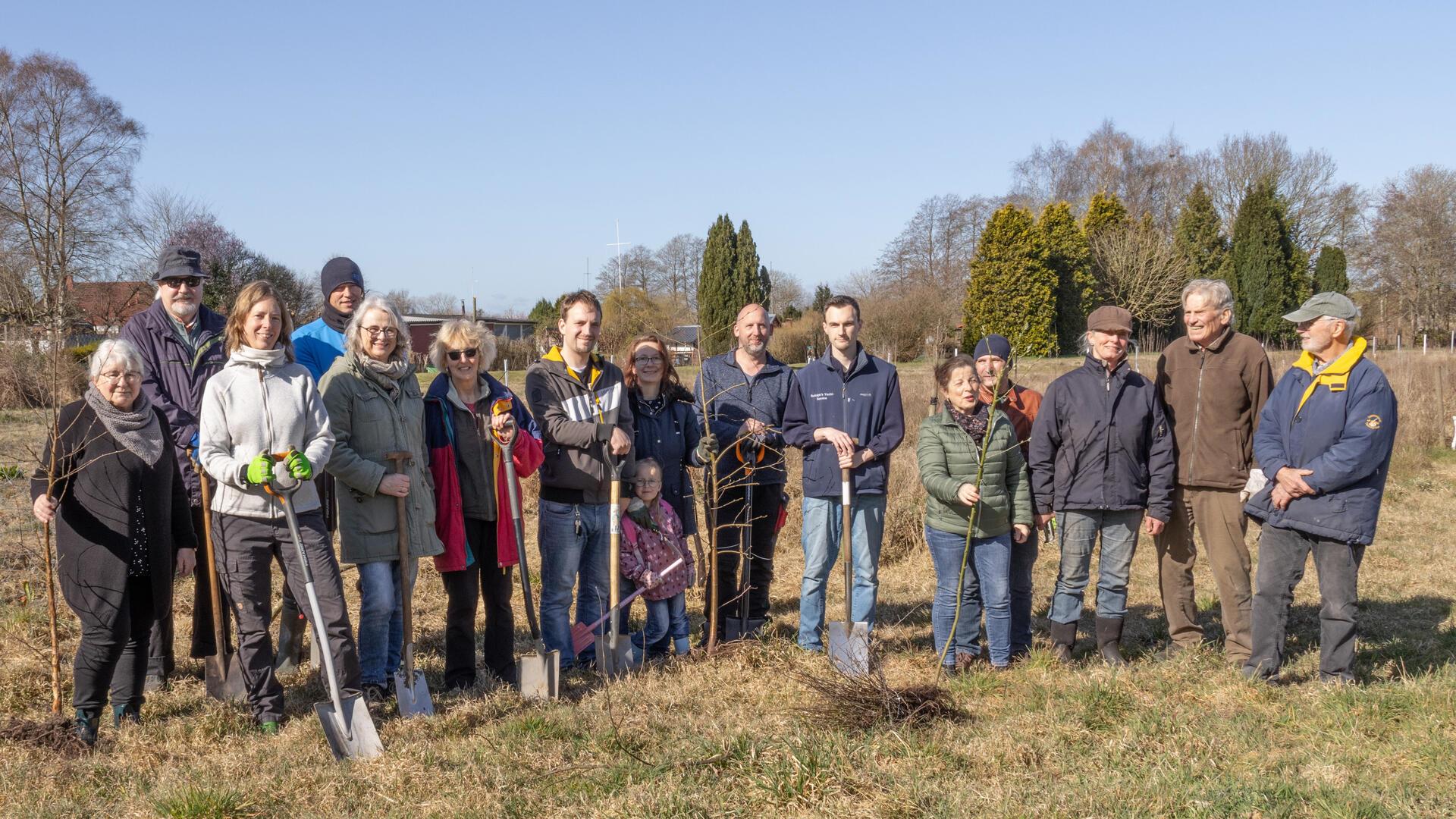 Eine Gruppe von 16 Menschen – von Kindern bis Senioren – steht auf einer Wiese an einem sonnigen Tag. Sie halten Schaufeln und junge Bäume.
