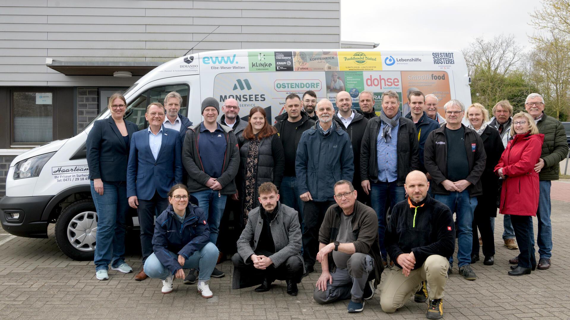 Gruppenfoto von rund 25 Personen vor einem weißen Transporter mit zahlreichen Sponsorenlogos; die Gruppe steht lächelnd auf einem Parkplatz vor einem modernen Gebäude.