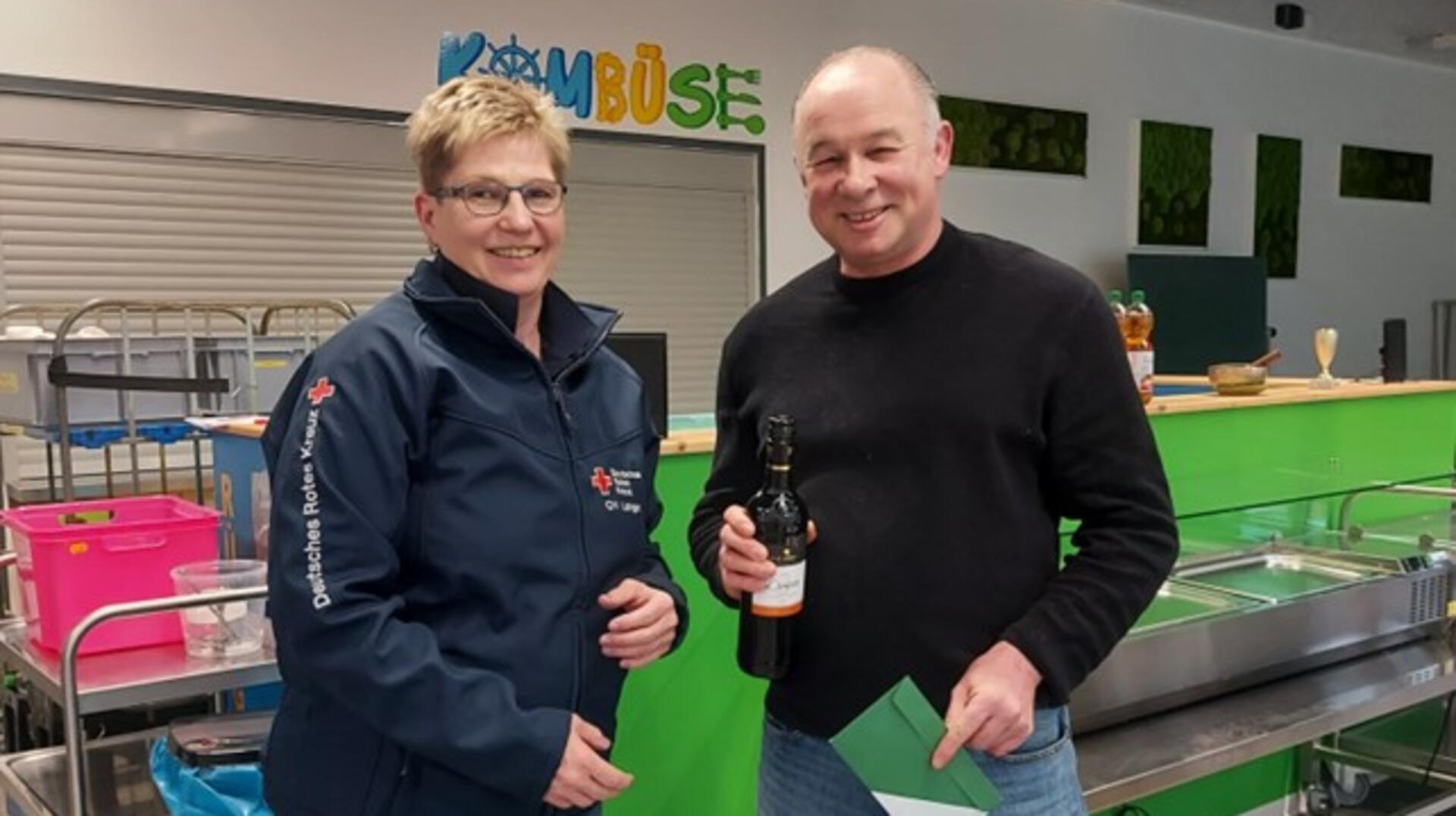 Eine Frau in DRK-Jacke und ein Mann mit Weinflasche und Umschlag stehen lächelnd in einer Schulküche vor einer grünen Essensausgabe mit dem Schriftzug „Kombüse“.