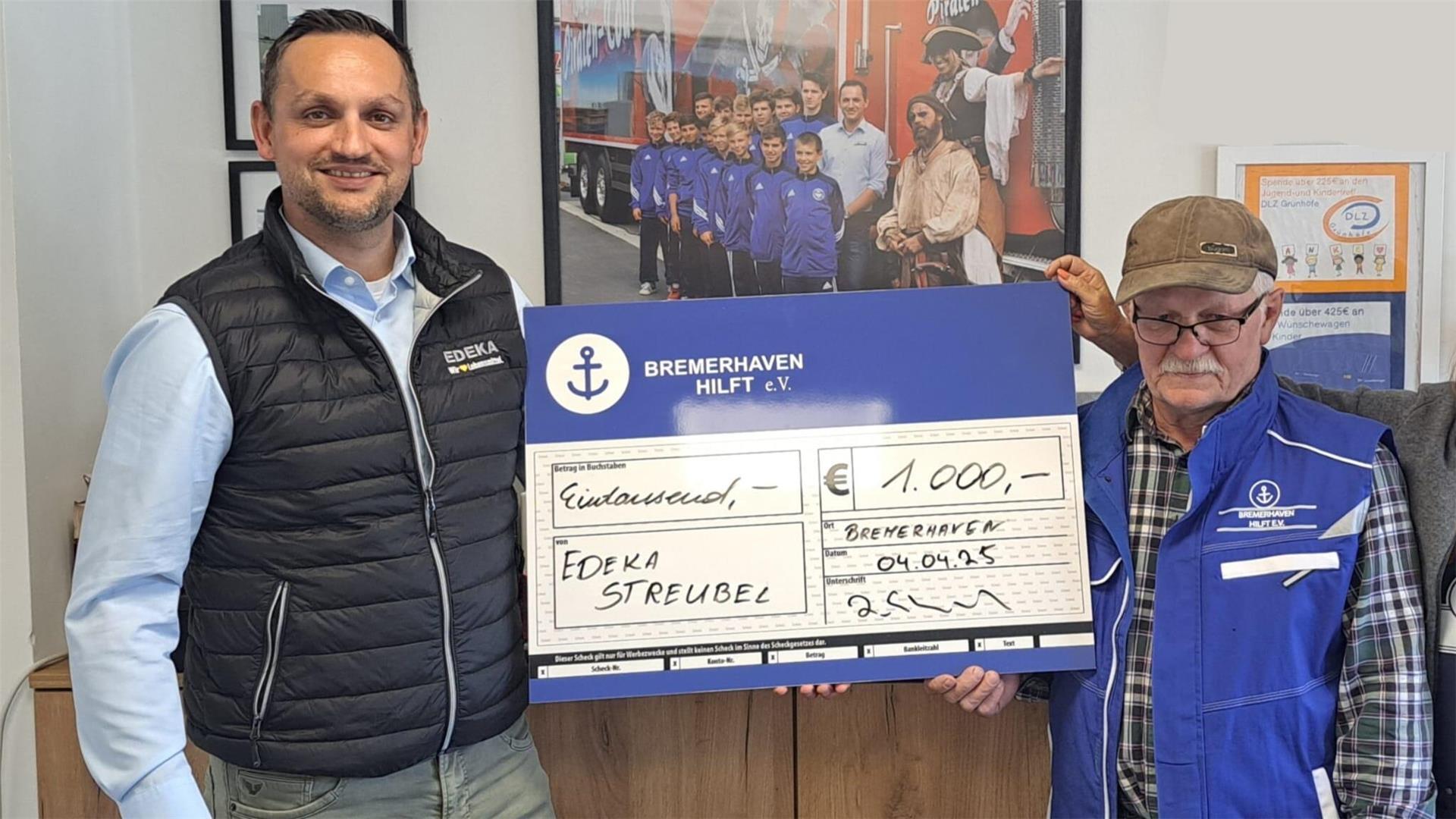 Zwei Männer stehen nebeneinander und halten gemeinsam einen überdimensionalen blauen Scheck über 1.000 Euro zugunsten von „Bremerhaven Hilft e.V.“, im Hintergrund hängt ein Bild von Jugendfußballern.