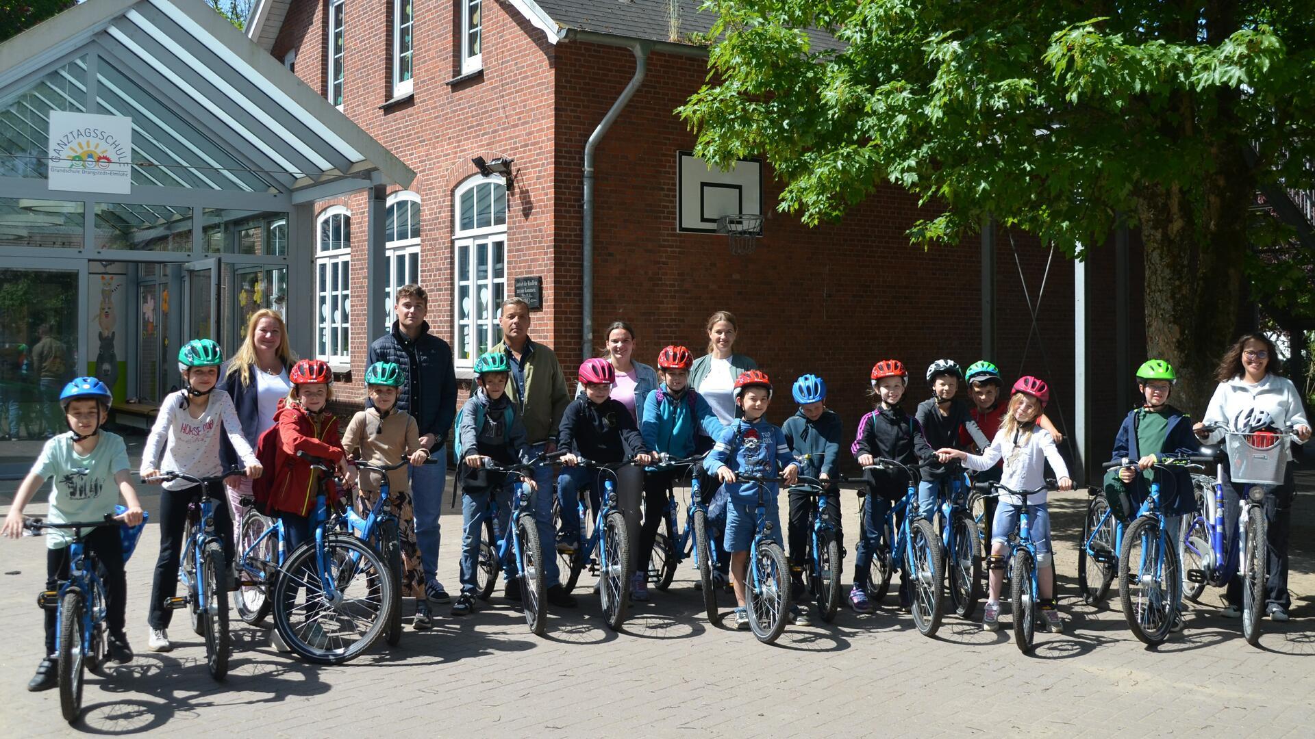 Eine Gruppe von Grundschulkindern mit Fahrradhelmen steht mit neuen Fahrrädern vor der Grundschule Drangstedt-Elmlohe, begleitet von mehreren Erwachsenen.