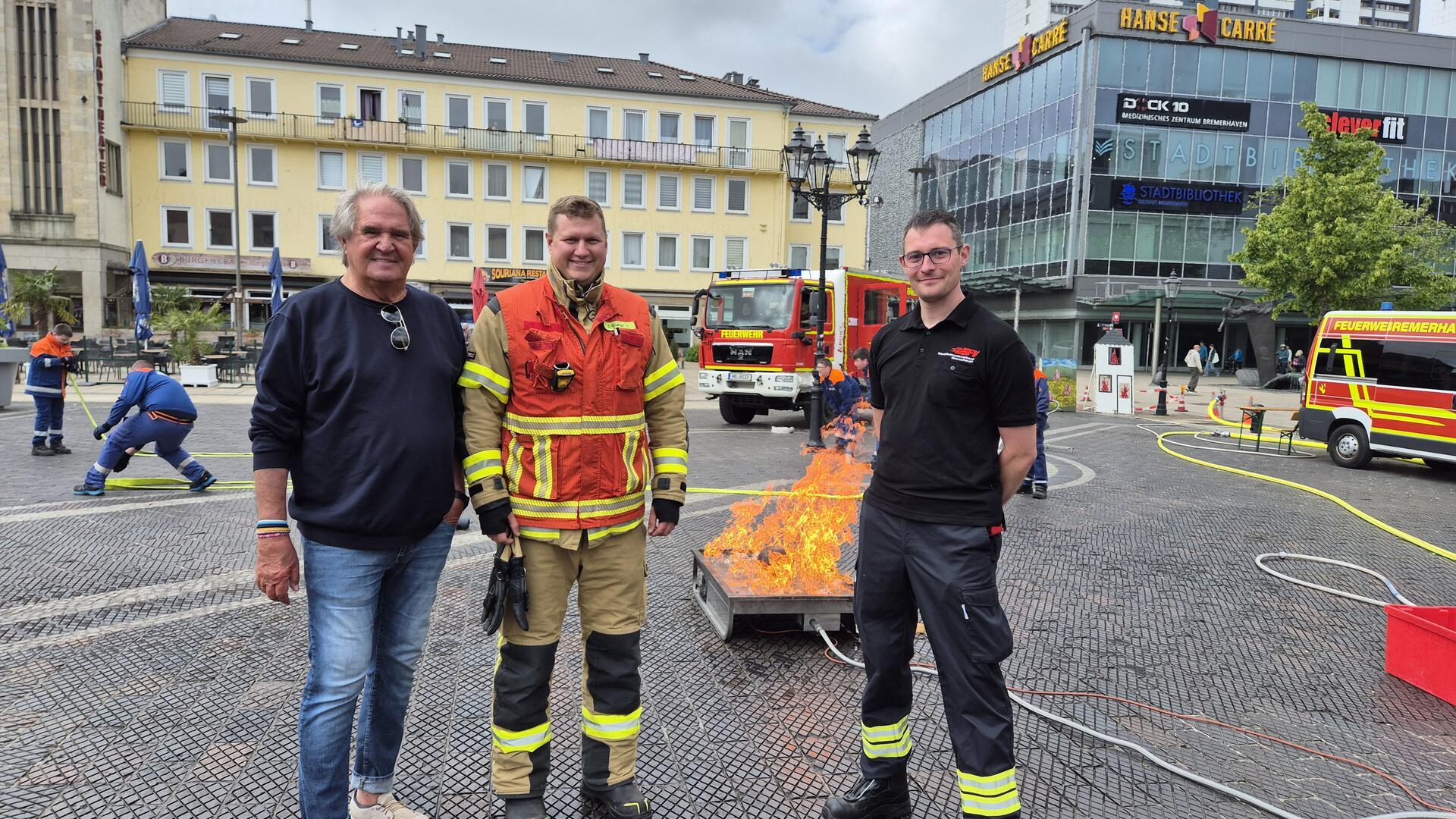 Drei Männer stehen nebeneinander. Im Hintergrund sind Flammen, ein Feuerwehrfahrzeug und ein Rettungswagen zu sehen.   