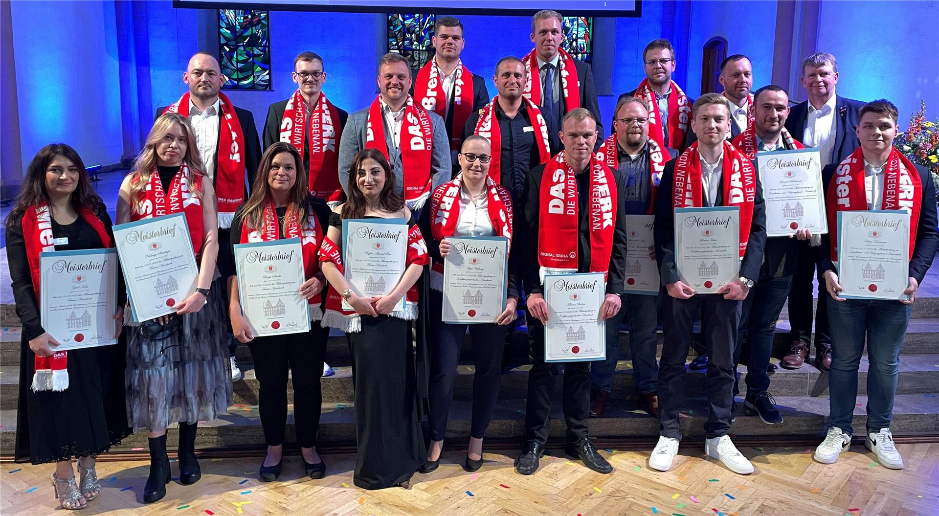 Gruppenfoto von rund 20 frischgebackenen Handwerksmeistern und -meisterinnen bei der Meisterfeier in Bremen. Die Personen stehen auf einer Bühne vor blau beleuchtetem Hintergrund und tragen rote Schals mit der Aufschrift „DAS HANDWERK – DIE WIRTSCHAFTSMACHT VON NEBENAN.“ Alle halten stolz ihre Meisterbriefe in den Händen. 