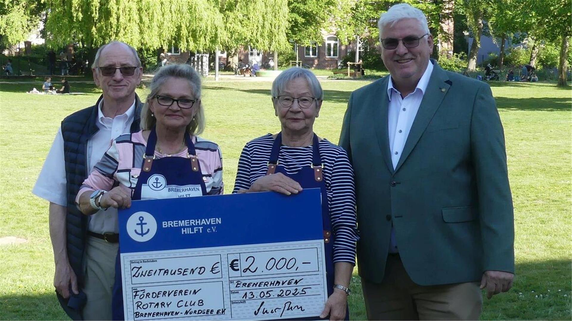 Vier Personen stehen im Freien auf einer Wiese, zwei davon halten einen großen symbolischen Spendenscheck über 2.000 Euro für den Verein "Bremerhaven hilft e.V." vom Förderverein Rotary Club Bremerhaven-Nordsee.