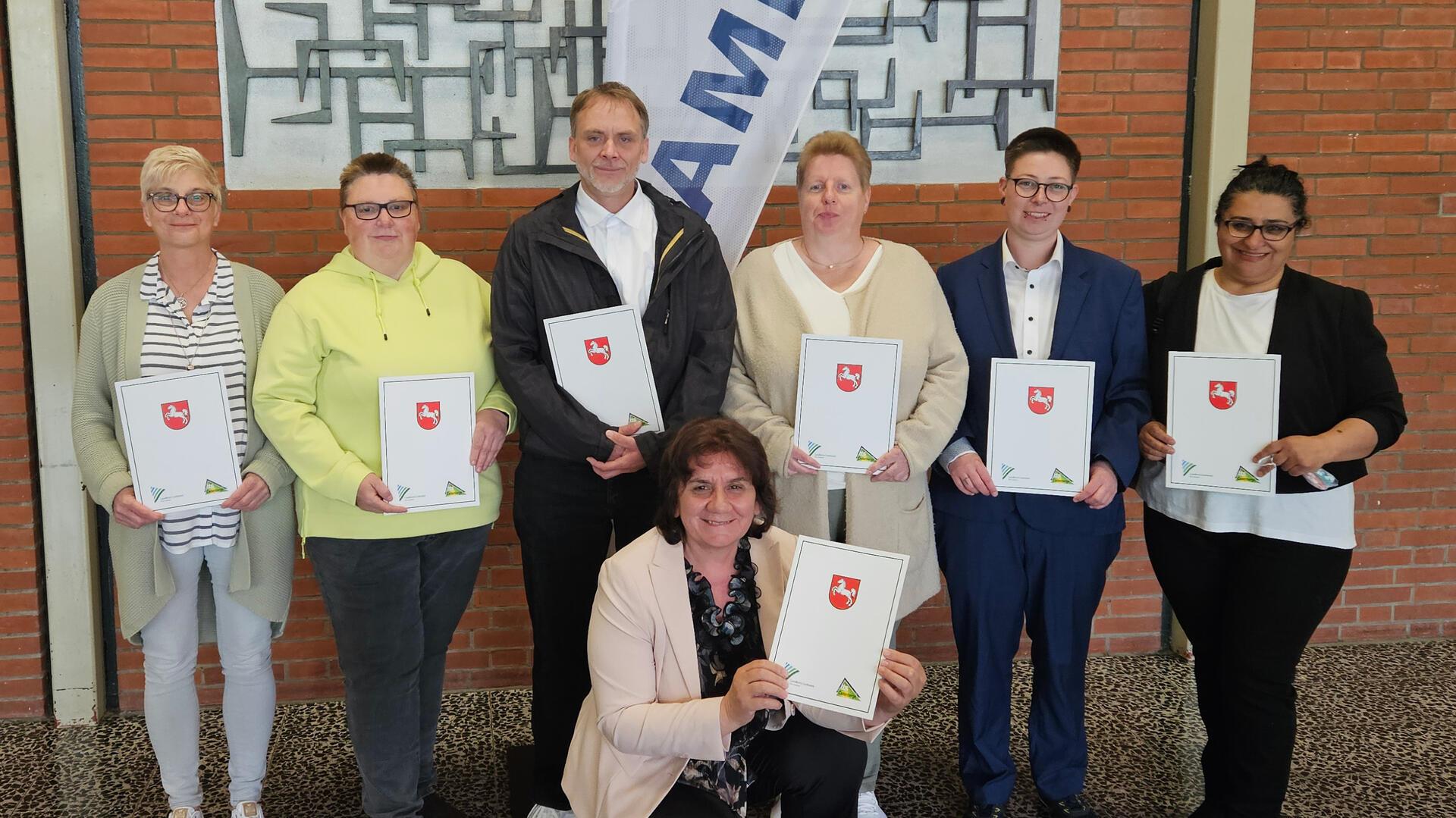 Sieben Mitarbeitende des AMEOS Klinikums Bremerhaven posieren mit ihren Abschlussurkunden des Kurzbildungsgangs Pflegeassistenz vor einem Werbewimpel mit AMEOS-Logo.