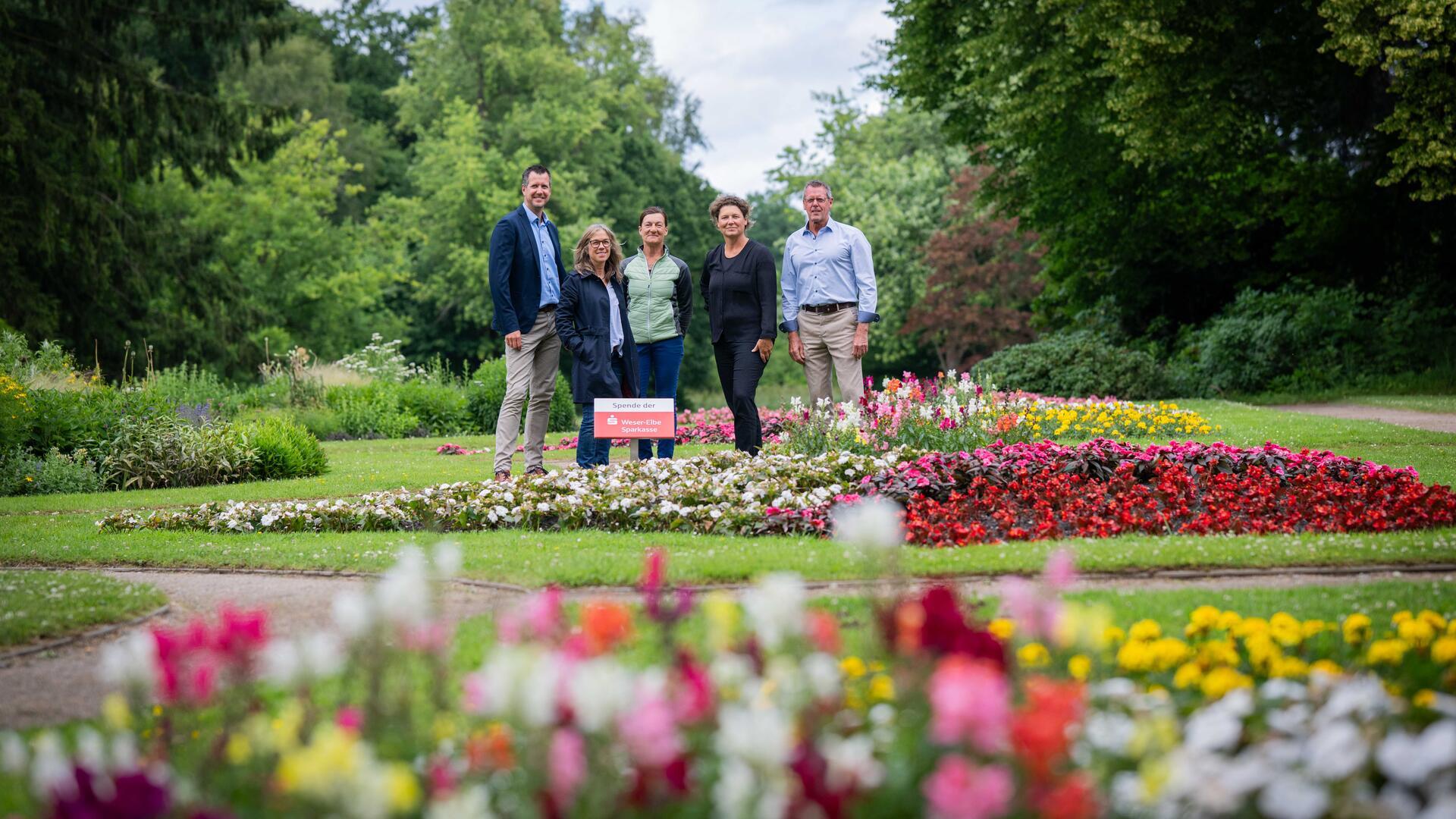 Fünf Personen stehen in einem bunt bepflanzten Blumenbeet im Bürgerpark. Im Vordergrund und Hintergrund blühen zahlreiche Sommerblumen in kräftigen Farben. Ein Schild weist auf eine Spende der Weser-Elbe Sparkasse hin. Die Szene ist von grünem Parkgelände und Bäumen umgeben.
