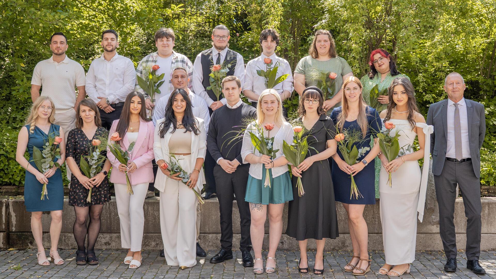 Gruppenfoto von 18 jungen Erwachsenen, die ihre Ausbildung zu Verwaltungsfachangestellten abgeschlossen haben. Sie stehen in zwei Reihen im Freien, halten Blumen in der Hand und lächeln in die Kamera. Rechts im Bild steht ein Mann im Anzug, vermutlich ein Vertreter des Magistrats. Im Hintergrund ist grünes Laub zu sehen, die Szene wirkt festlich und sommerlich.