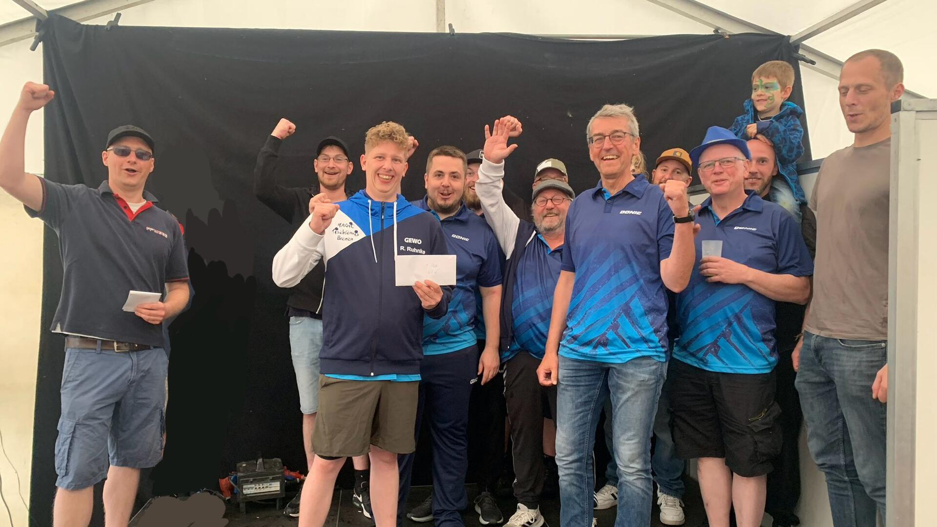 Gruppe jubelnder Männer, teils in blauen Sporttrikots, steht vor schwarzem Hintergrund in einem Festzelt. Zwei Personen halten Umschläge, ein Kind mit Gesichtsbemalung sitzt auf den Schultern.