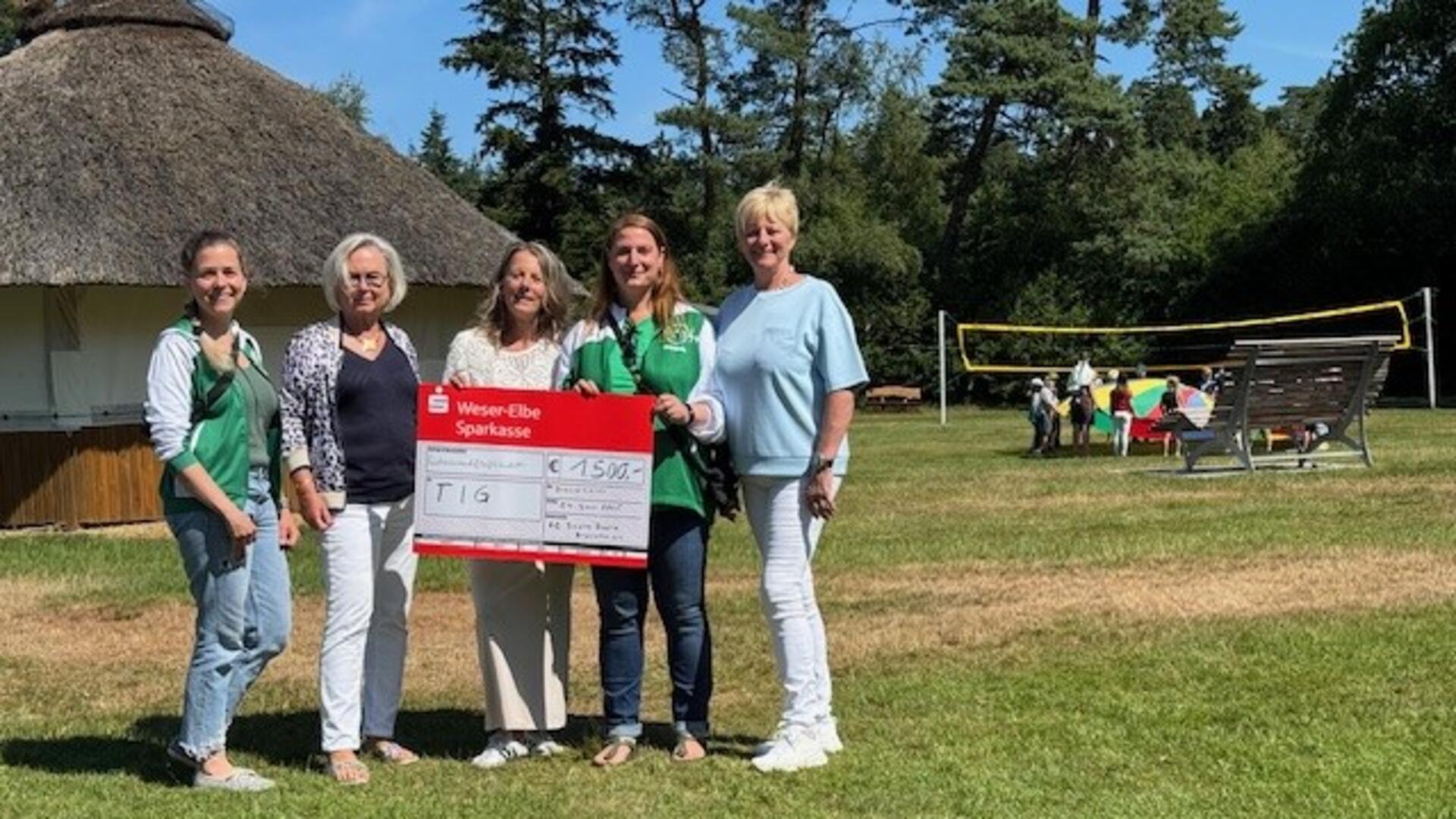 Fünf Frauen stehen auf einer Wiese vor einem runden Reetdachhaus und halten gemeinsam einen großen symbolischen Spendenscheck der Weser-Elbe Sparkasse über 1500 Euro. Im Hintergrund spielen Kinder mit einem bunten Schwungtuch auf einem Volleyballfeld.