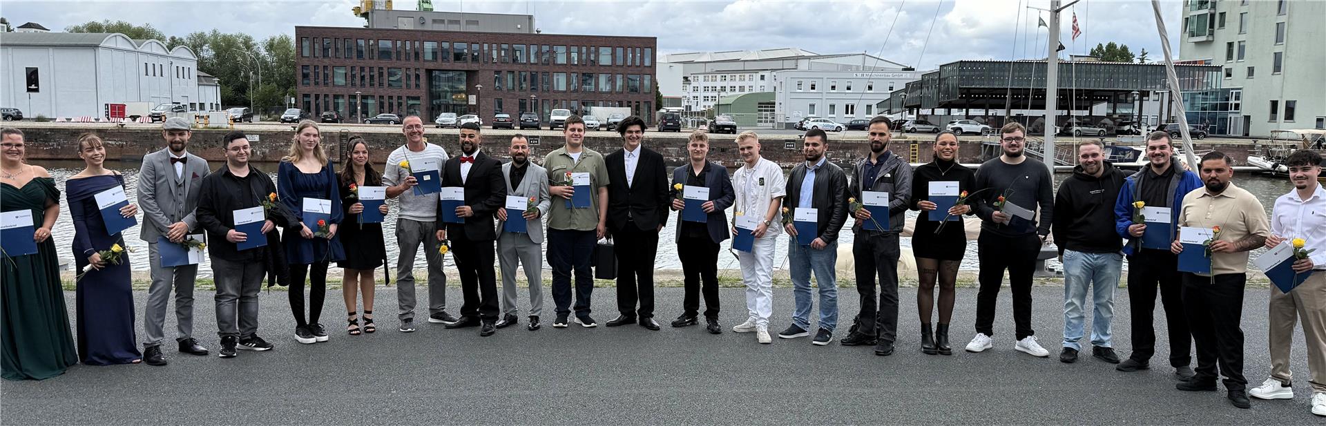 Gruppe junger Erwachsener steht in festlicher Kleidung am Hafen, viele halten Zeugnisse und einzelne Blumen in der Hand. Im Hintergrund sind Wasser, Boote und Gebäude zu sehen.