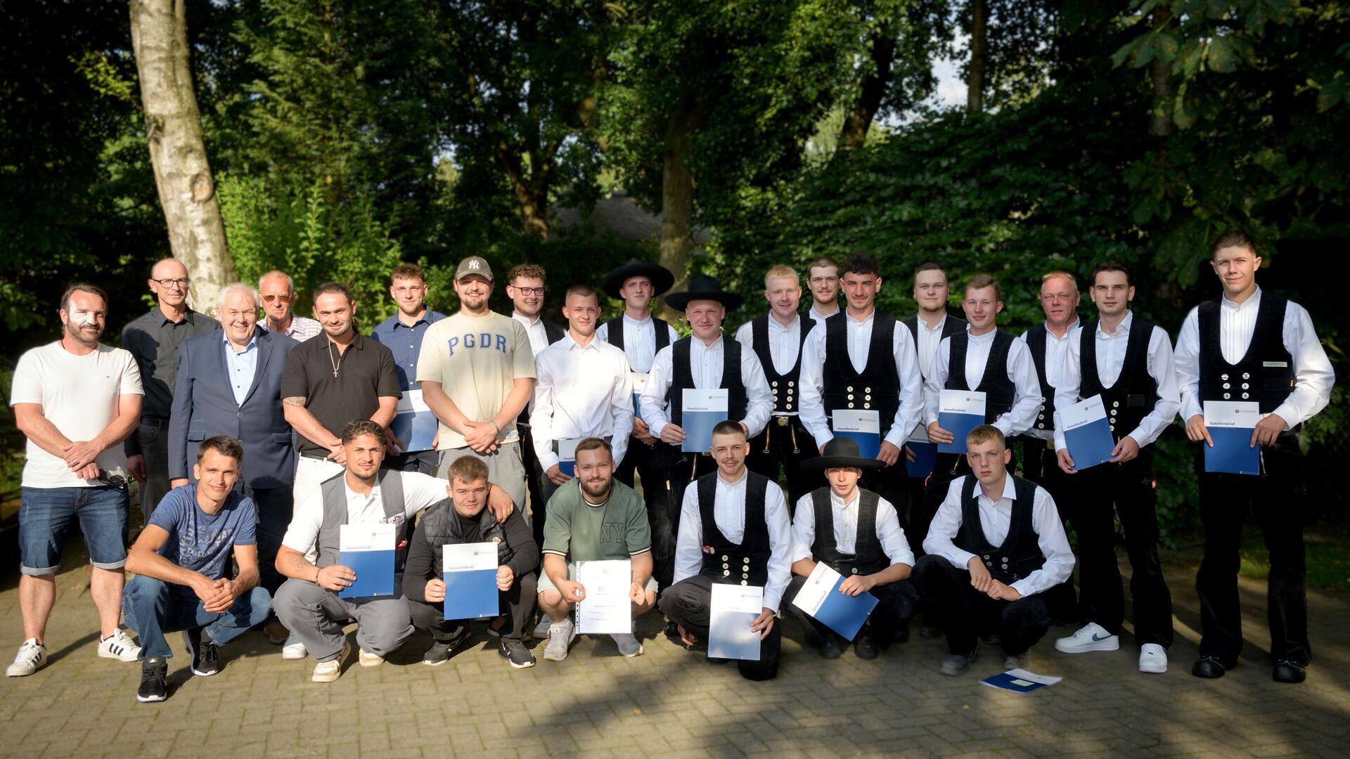 Gruppenfoto von jungen Männern, teils in traditioneller Handwerkskleidung, teils in Freizeit- oder festlicher Kleidung, viele mit Zeugnismappen in der Hand. Die Gruppe steht im Freien vor grünem, bewaldetem Hintergrund