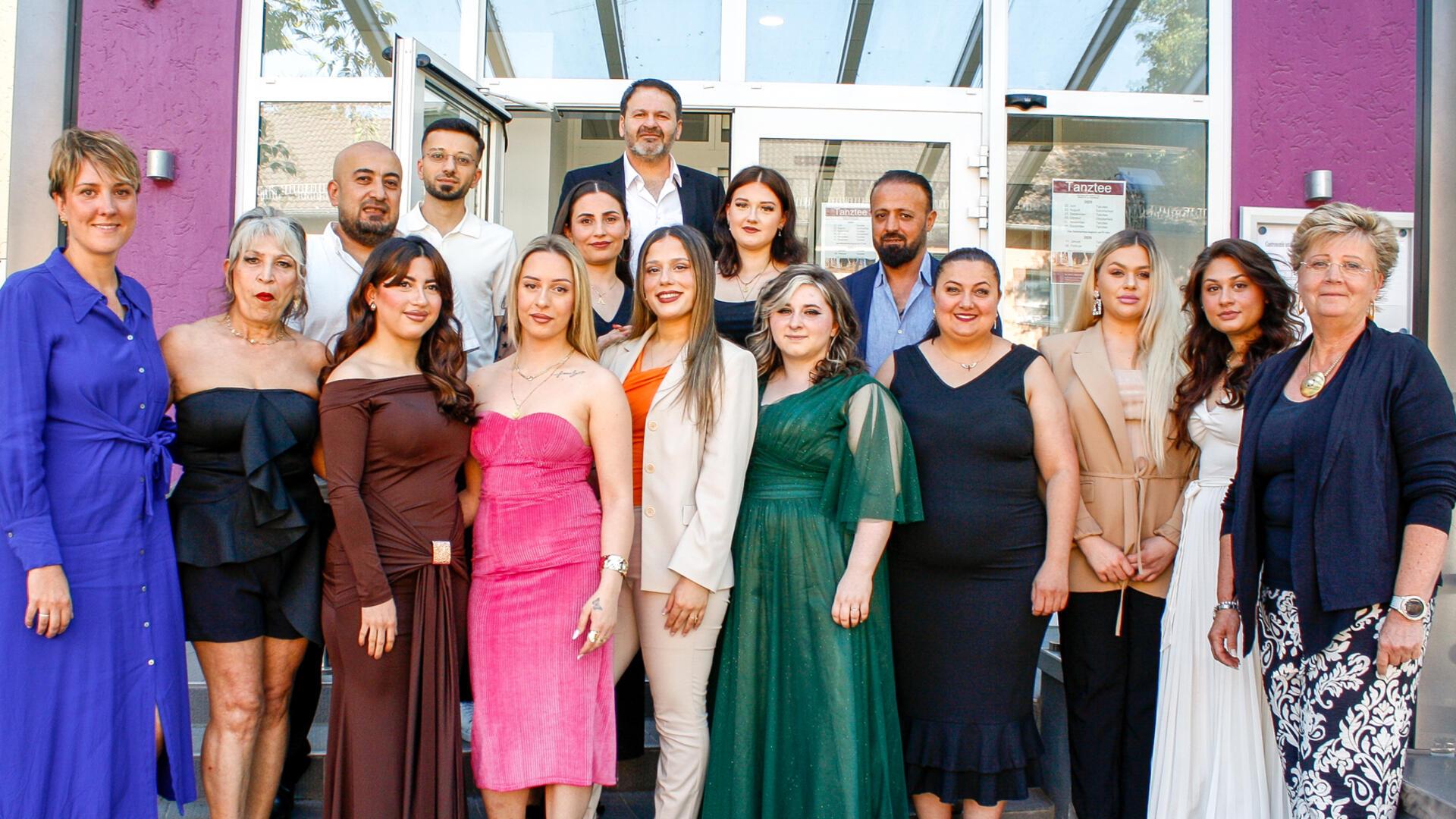 Gruppenfoto von 16 Personen in festlicher Kleidung vor einem Gebäude mit Glasdach und lilafarbener Wand. Männer und Frauen posieren gemeinsam für ein Bild.