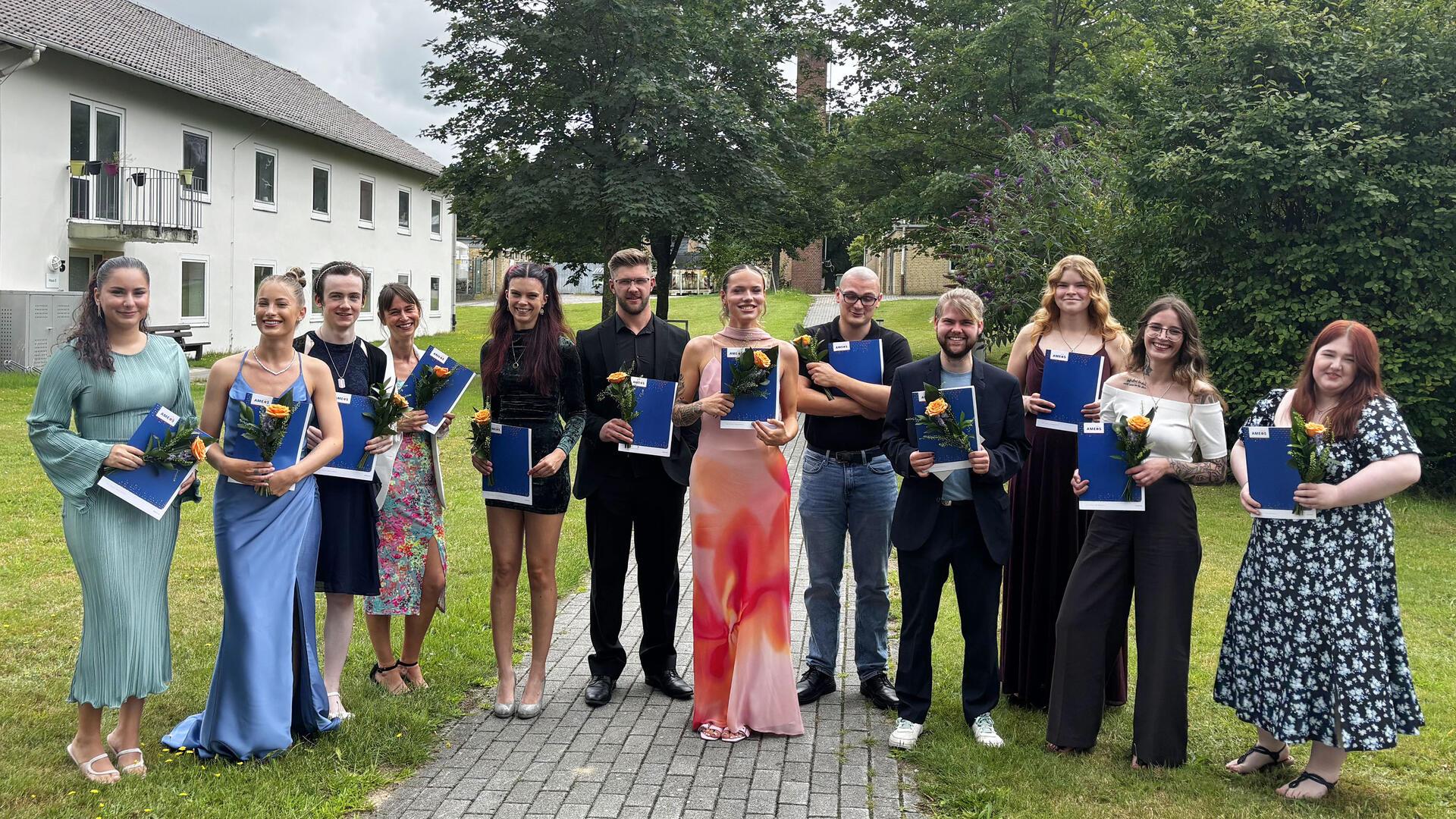 Zwölf frisch examinierte Pflegefachkräfte stehen mit Zeugnismappen und Blumen in der Hand lächelnd nebeneinander auf einem Weg im Grünen vor dem AMEOS Klinikum.