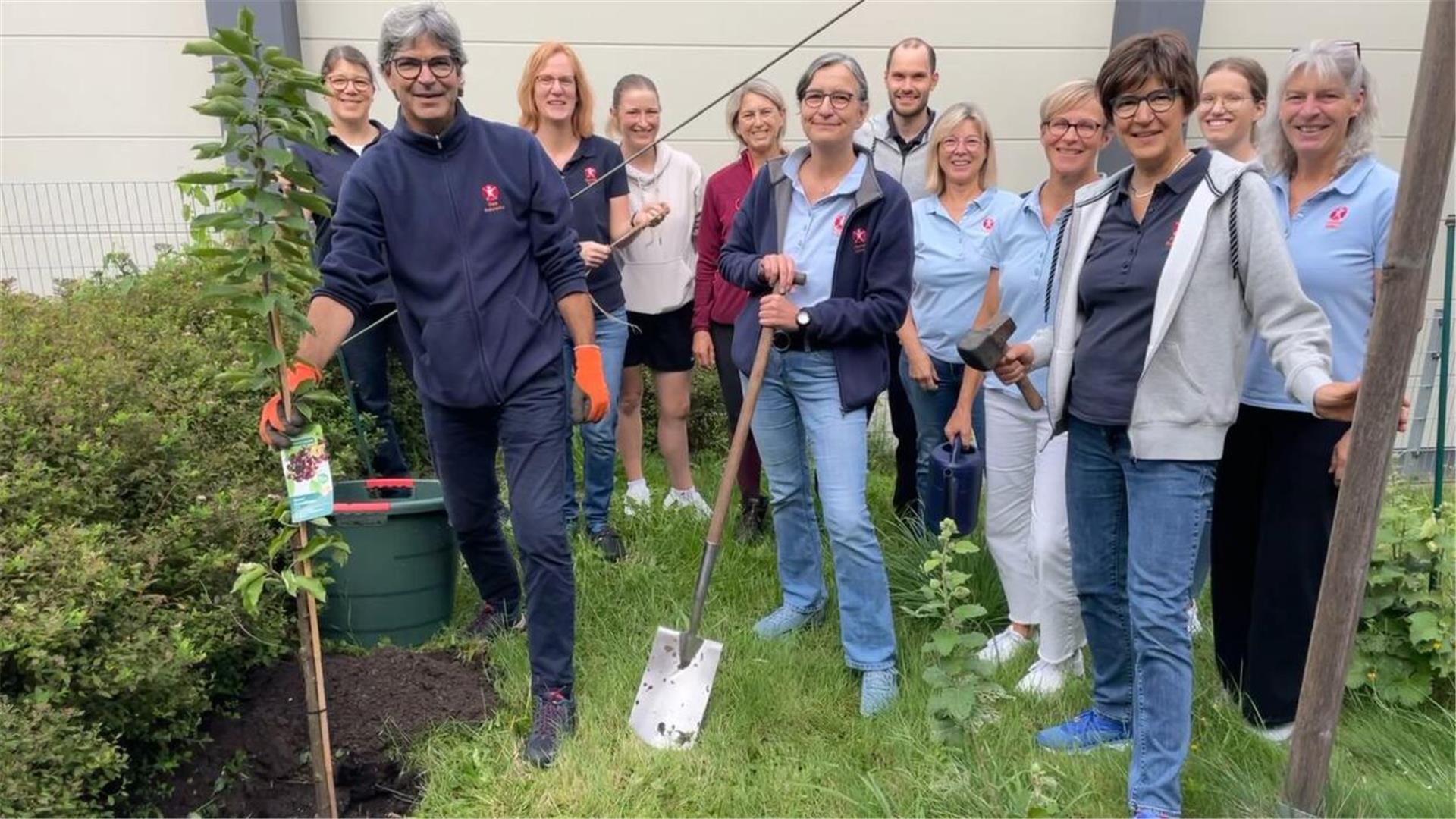 Eine Gruppe Männer und Frauen steht zusammen. Einige haben Werkzeug in der Hand und einer einen Baum.