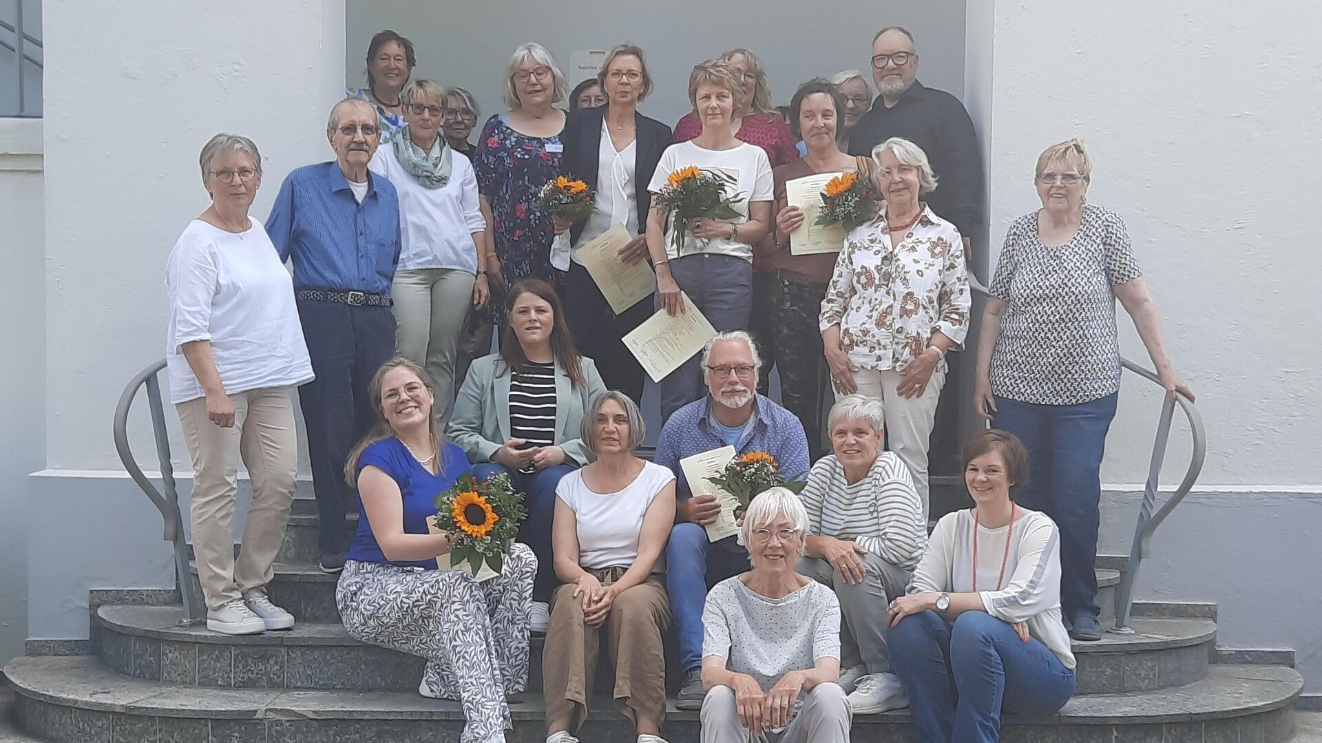 Menschen stehen vor einem Gebäude und halten Zertifikate und Blumen in der Hand. 