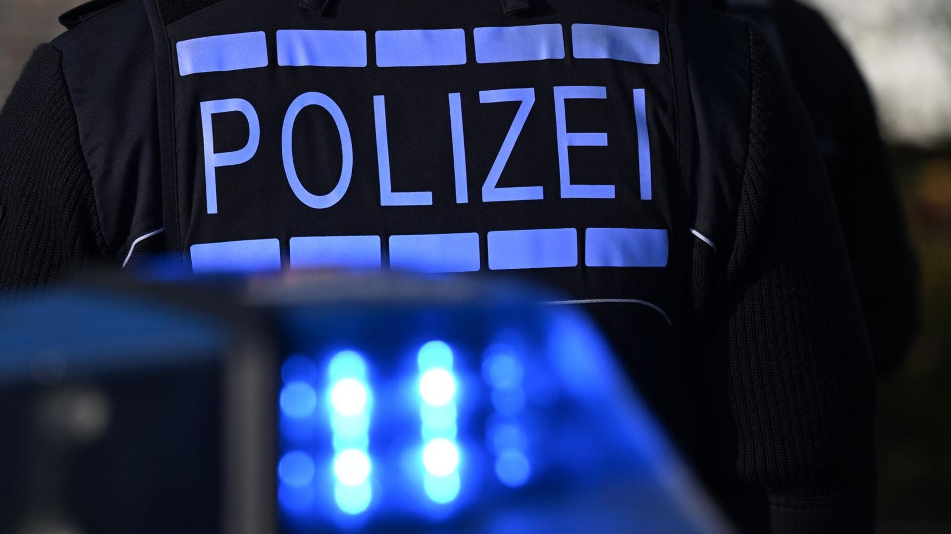 Das Logo der Polizei auf der Dienstkleidung eines Polizisten der vor einem Blaulicht steht 
