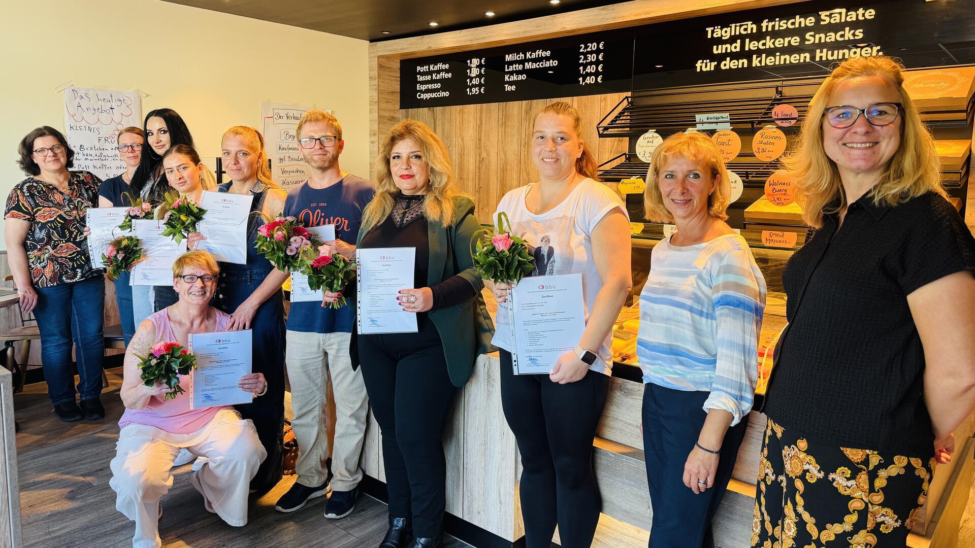 Eine Gruppe von Teilnehmerinnen und Teilnehmern einer BBU-Fortbildung im Lebensmittelverkauf steht in einer Bäckereifiliale. Mehrere Personen halten Zertifikate und Blumensträuße, flankiert von Mitarbeiterinnen des Projekts. Im Hintergrund sind eine Verkaufstheke mit Snacks sowie eine Preistafel zu sehen.
