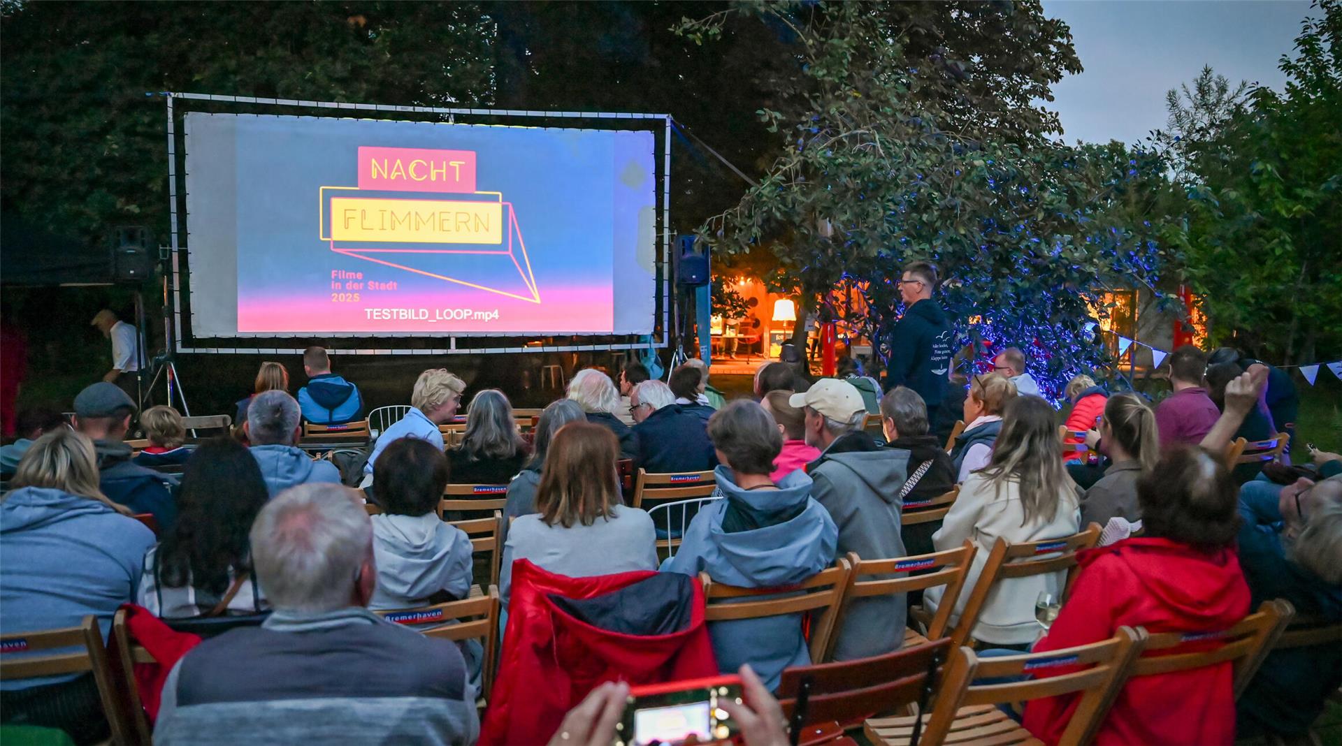 Menschen sitzen auf Stühlen vor einer Leinwand im Freien.