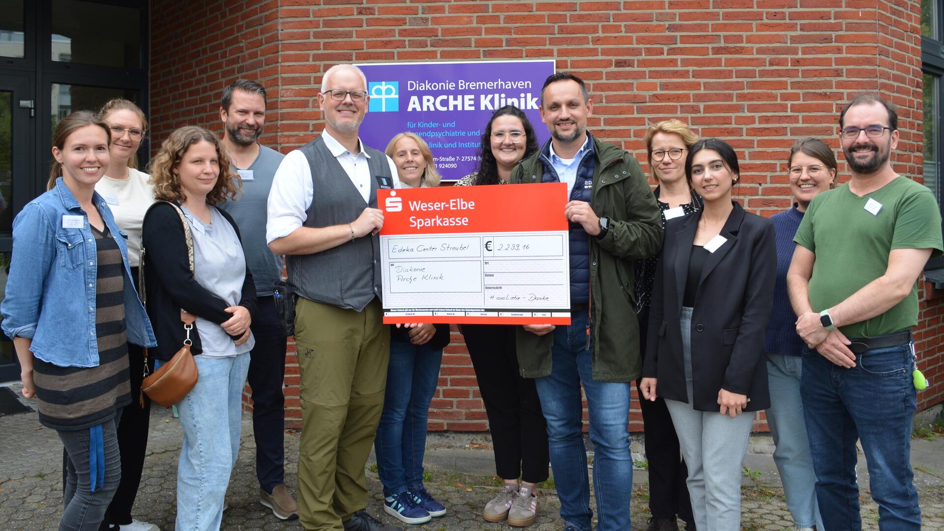 Mitarbeitende der Diakonie Bremerhaven und Vertreter des Edeka Centers stehen gemeinsam vor der ARCHE Klinik und präsentieren einen symbolischen Spendenscheck über 2.239,16 Euro.