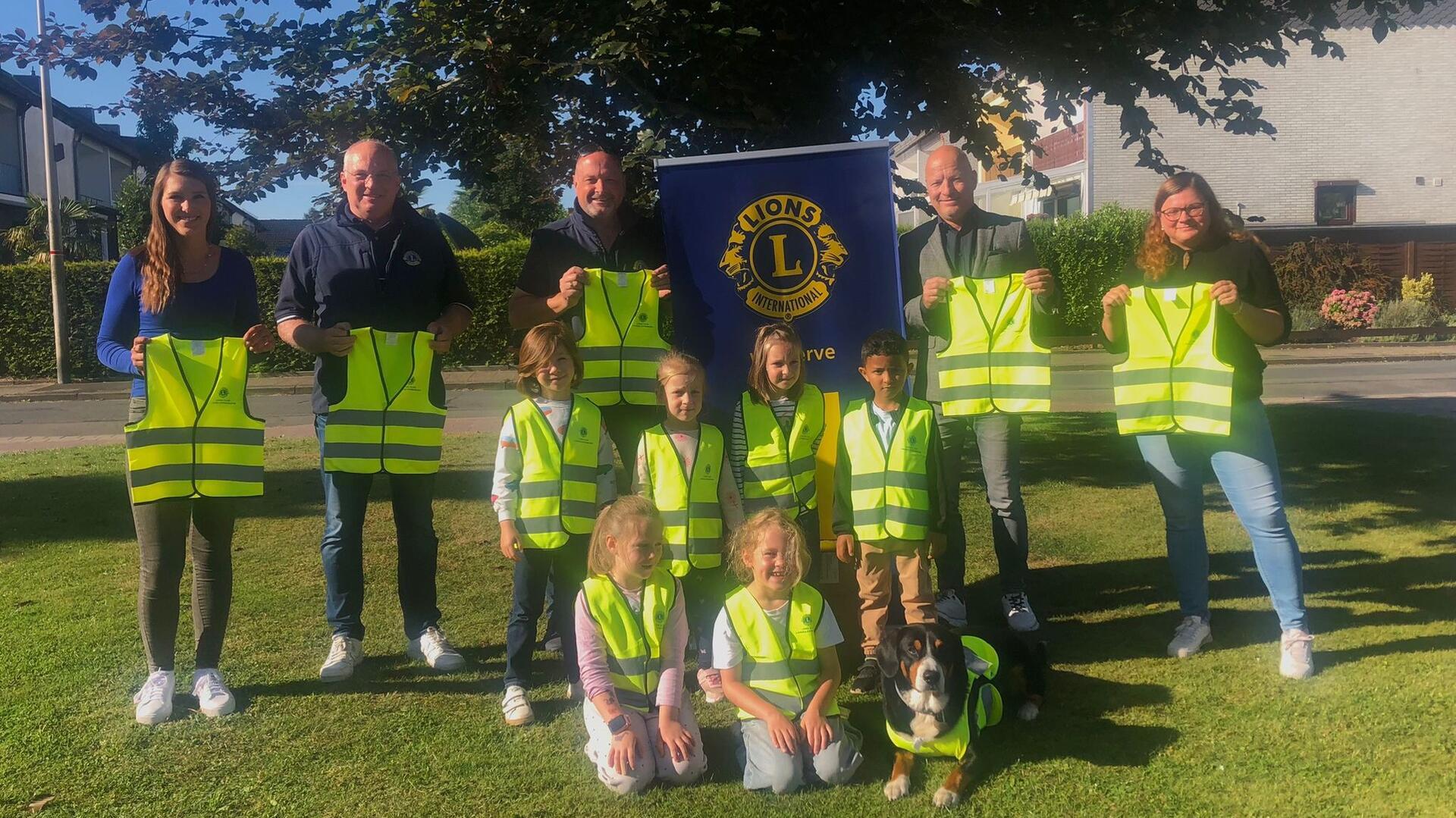 Mehrere Erwachsene und Kinder stehen und knien auf einer Wiese, alle tragen oder halten leuchtend gelbe Sicherheitswesten. Hinter ihnen ist ein Banner des Lions Clubs zu sehen. Auch ein Hund trägt eine Warnweste.