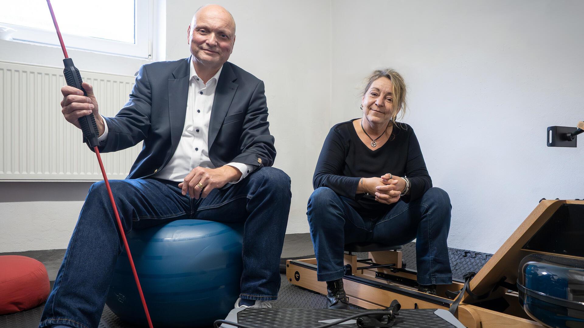Ein Mann im Anzug sitzt auf einem blauen Gymnastikball und hält ein Trainingsgerät mit rotem Widerstandsband. Neben ihm sitzt eine Frau in schwarzem Oberteil und Jeans auf einem Sportgerät.