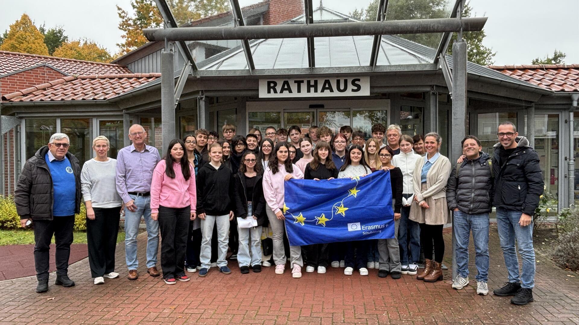 Eine große Gruppe von Schülerinnen, Schülern und Lehrkräften steht vor dem Loxstedter Rathaus. In der ersten Reihe halten einige eine blaue Erasmus+-Flagge mit gelben Sternen.