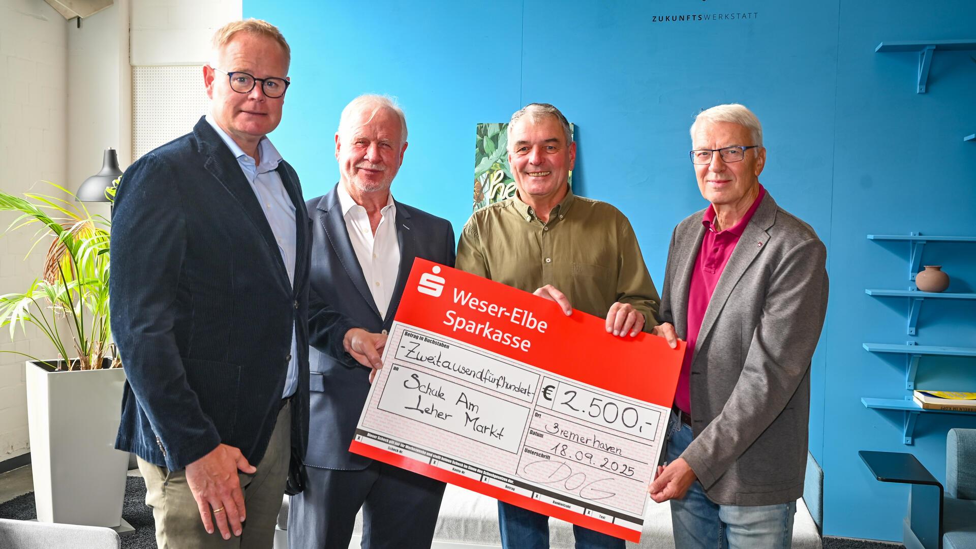 Vier Männer stehen nebeneinander und halten gemeinsam einen großen symbolischen Spendenscheck über 2.500 Euro.