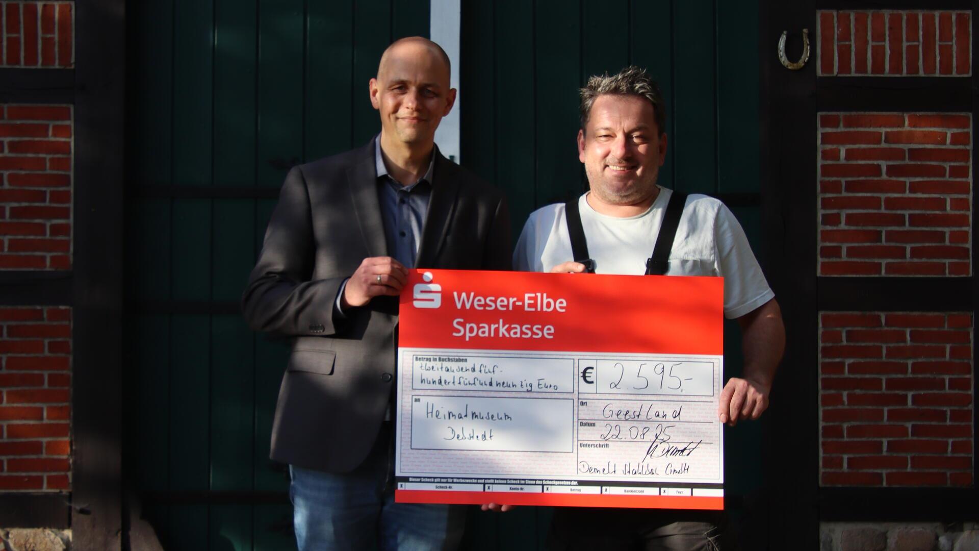 Zwei Männer stehen vor einem grünen Tor mit Fachwerkrahmen und halten gemeinsam einen großen symbolischen Spendenscheck der Weser-Elbe Sparkasse. Der Scheck ist auf das Heimatmuseum Debstedt ausgestellt und zeigt eine Spendensumme von 2.595 Euro.