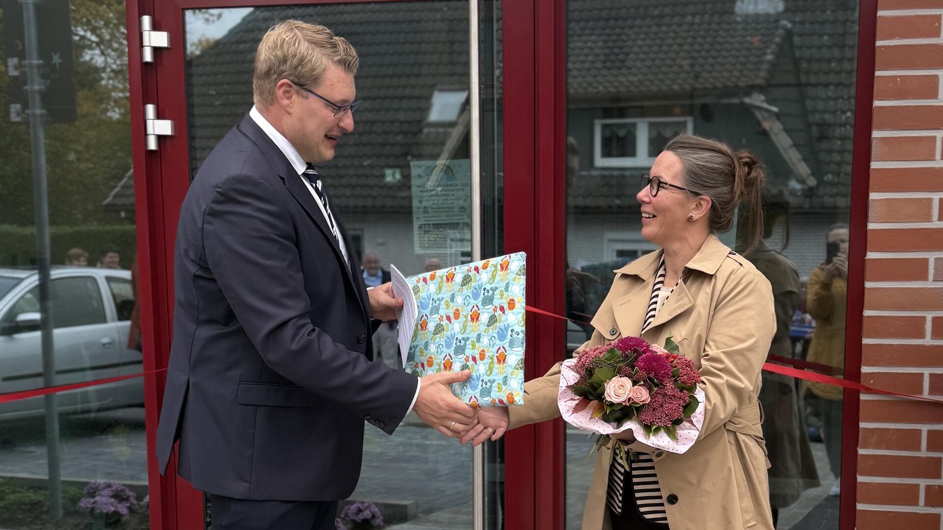Ein Mann im Anzug überreicht einer Frau mit Blumenstrauß vor einem Gebäude ein Geschenk. Beide lächeln, im Hintergrund ist ein rotes Band zur Eröffnung zu sehen.