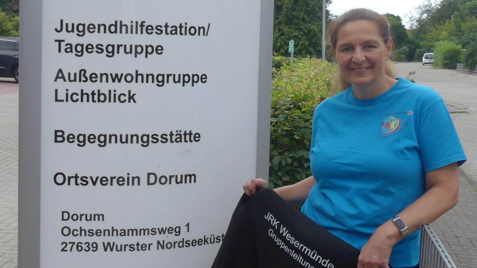 Frau in blauem Jugendrotkreuz-T-Shirt steht lächelnd neben einem Schild des DRK-Ortsvereins Dorum mit Adressangabe und Hinweisen auf soziale Einrichtungen.