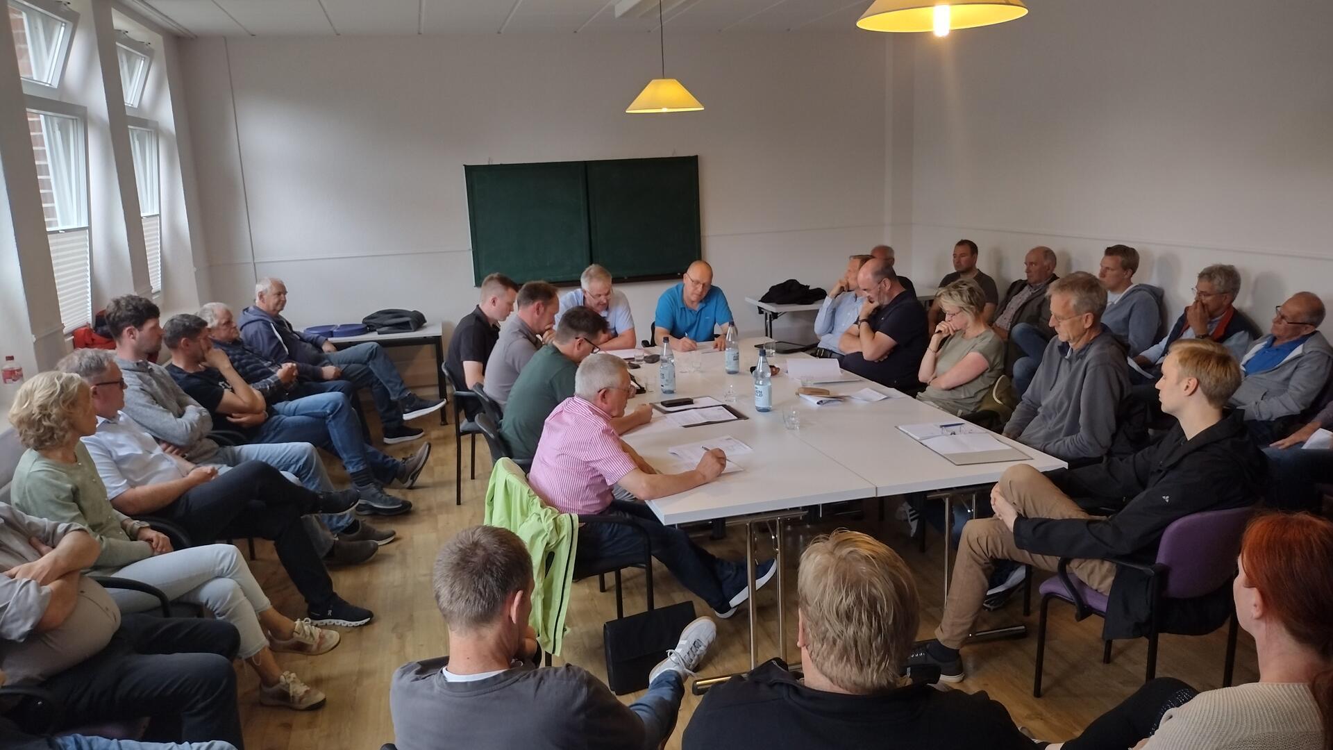 In der Bildmitte sitzen elf Menschen um einen Tisch herum, und an den Wänden sitzen Besucher auf langen Stuhlreihen.