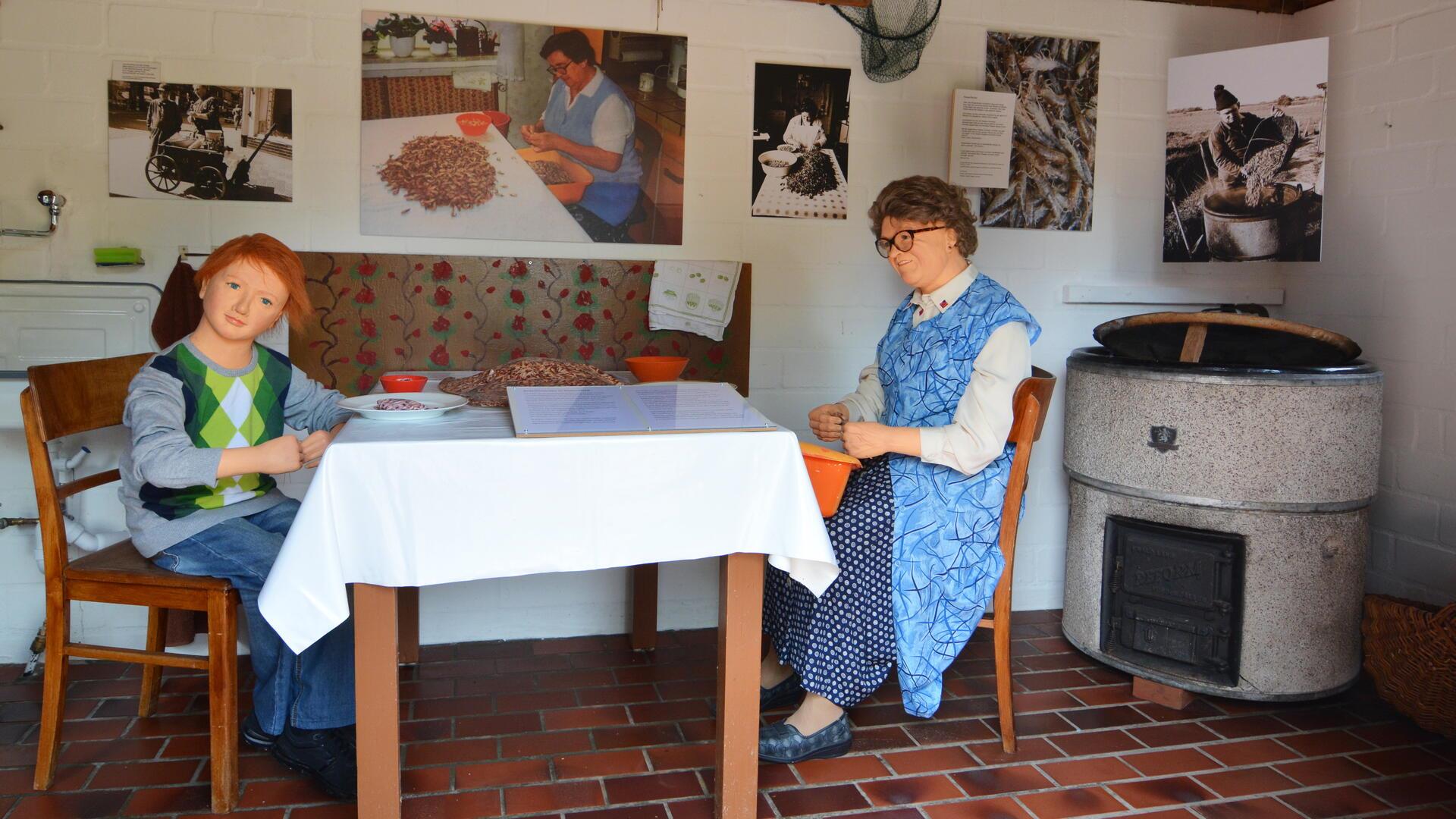 Blick ins Museum: Zwei Schaufensterpuppen sitzen auf Holzstühlen an einem Tisch. An der Wand hängen Fotos.