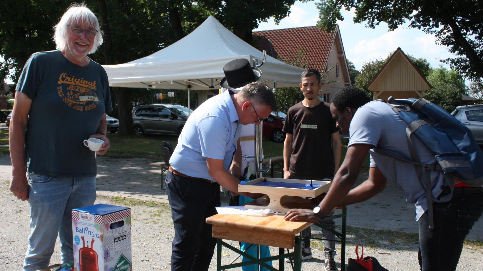Vier Männer stehen an einem Tisch im Freien, zwei von ihnen spielen ein Tischspiel. Einer trägt einen Rucksack, ein anderer eine helle Hemdbluse und lehnt sich über das Spiel. Im Hintergrund stehen ein weißes Pavillonzelt, parkende Autos und Häuser.