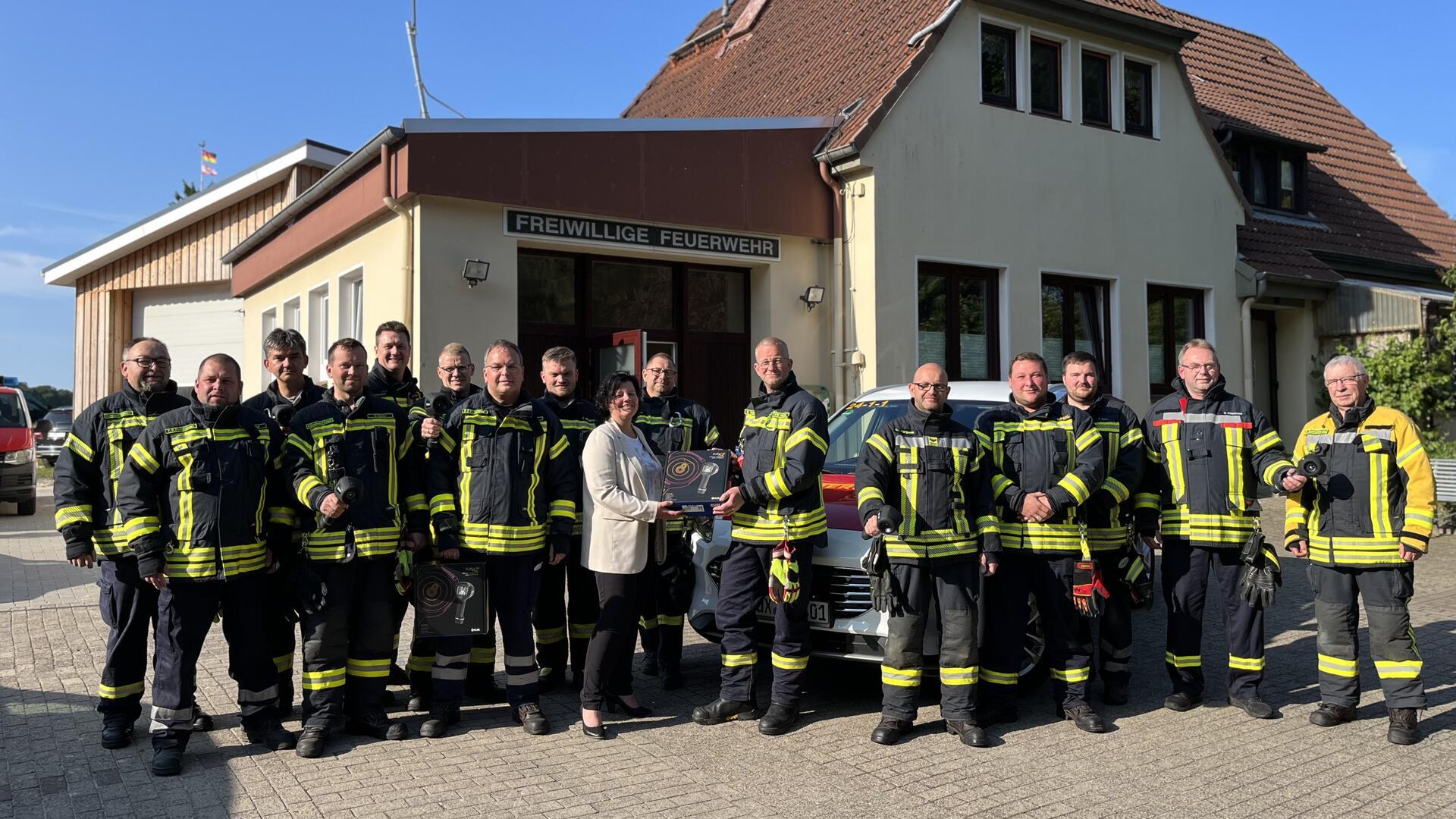 Gruppenfoto vor dem Gebäude der Freiwilligen Feuerwehr: Eine Gruppe von Feuerwehrleuten in Einsatzkleidung steht gemeinsam mit Bürgermeisterin Gabi Kasten, die eine Wärmebildkamera überreicht. Im Hintergrund sind Feuerwehrfahrzeuge und das Feuerwehrhaus zu sehen.