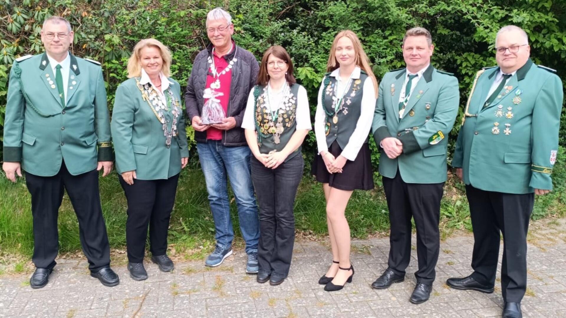 Gruppenfoto von sieben Mitgliedern eines Schützenvereins in Uniform oder mit Schützenorden, aufgenommen im Freien vor grüner Hecke. Eine Person hält ein Geschenk.
