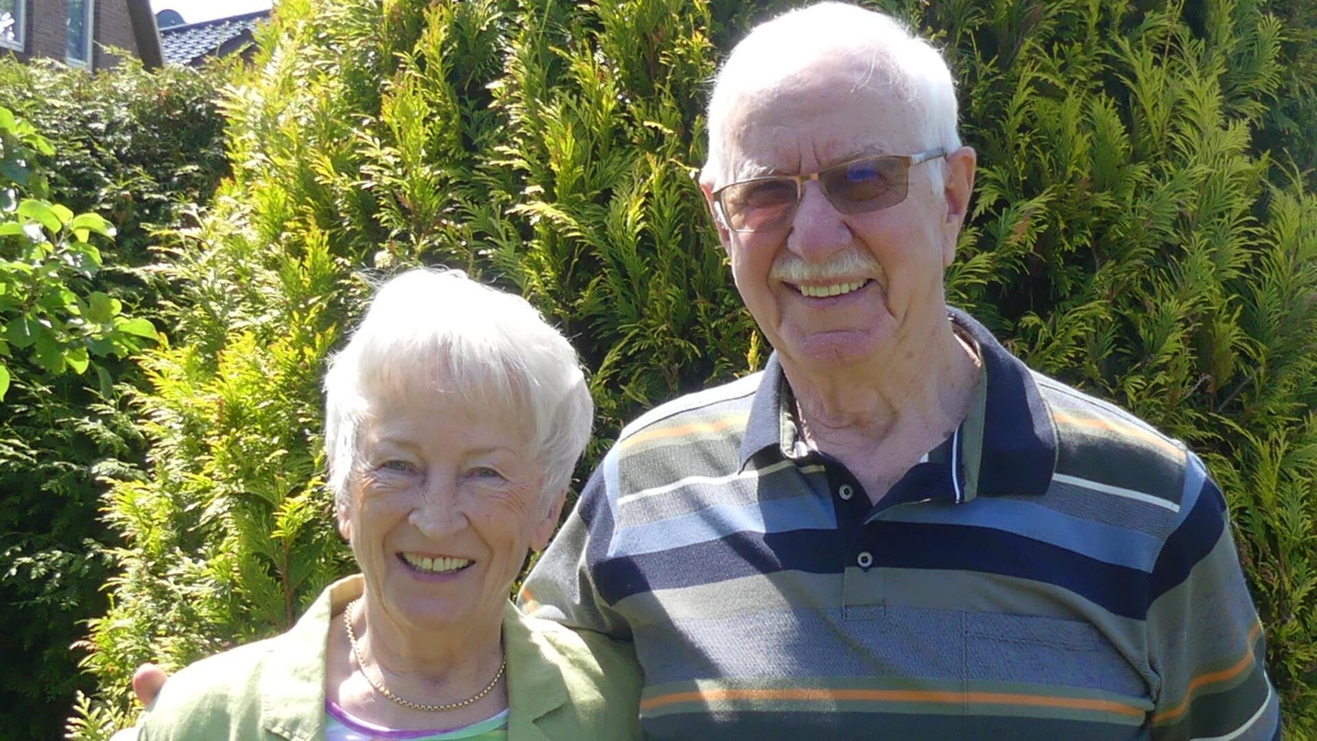 Älteres Ehepaar steht lächelnd nebeneinander vor einer Hecke im Garten; beide tragen sommerliche Kleidung, die Frau eine helle, dünne Jacke, der Mann ein gestreiftes Poloshirt.