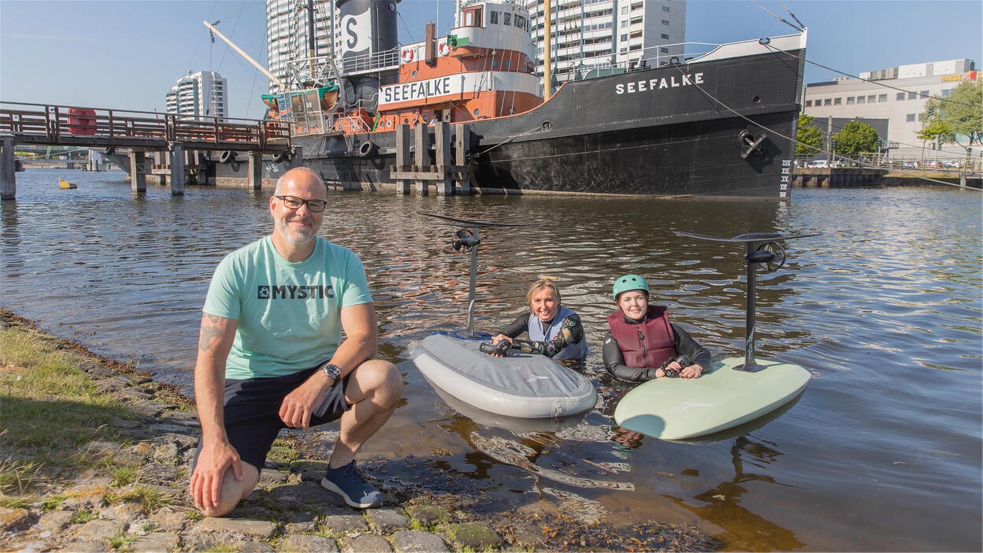 Drei Personen posieren für ein Foto. Zwei davon sind mit Surfboards im Wasser.