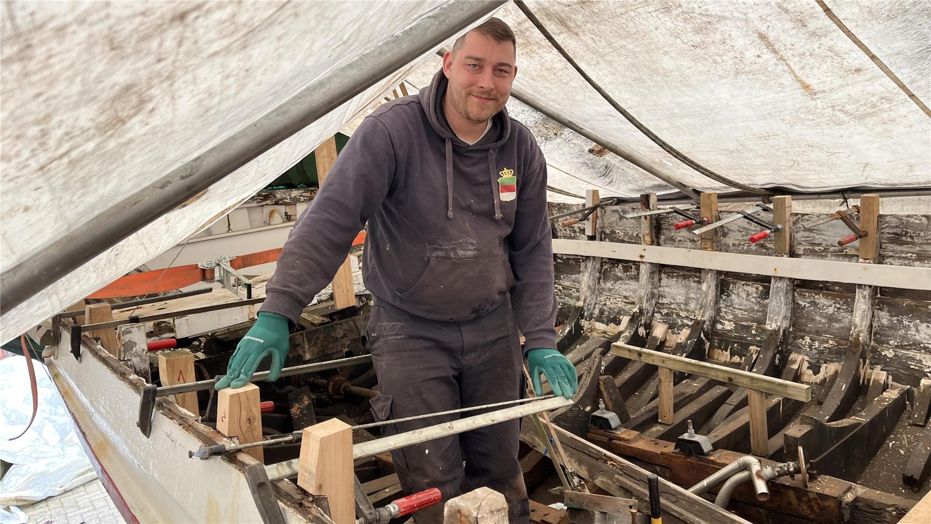 Mann steht auf einem Boot unter einer Plane. Christian Schwan macht ein Börteboot wieder fit. Zur Sail im August soll es spätestens fahren.