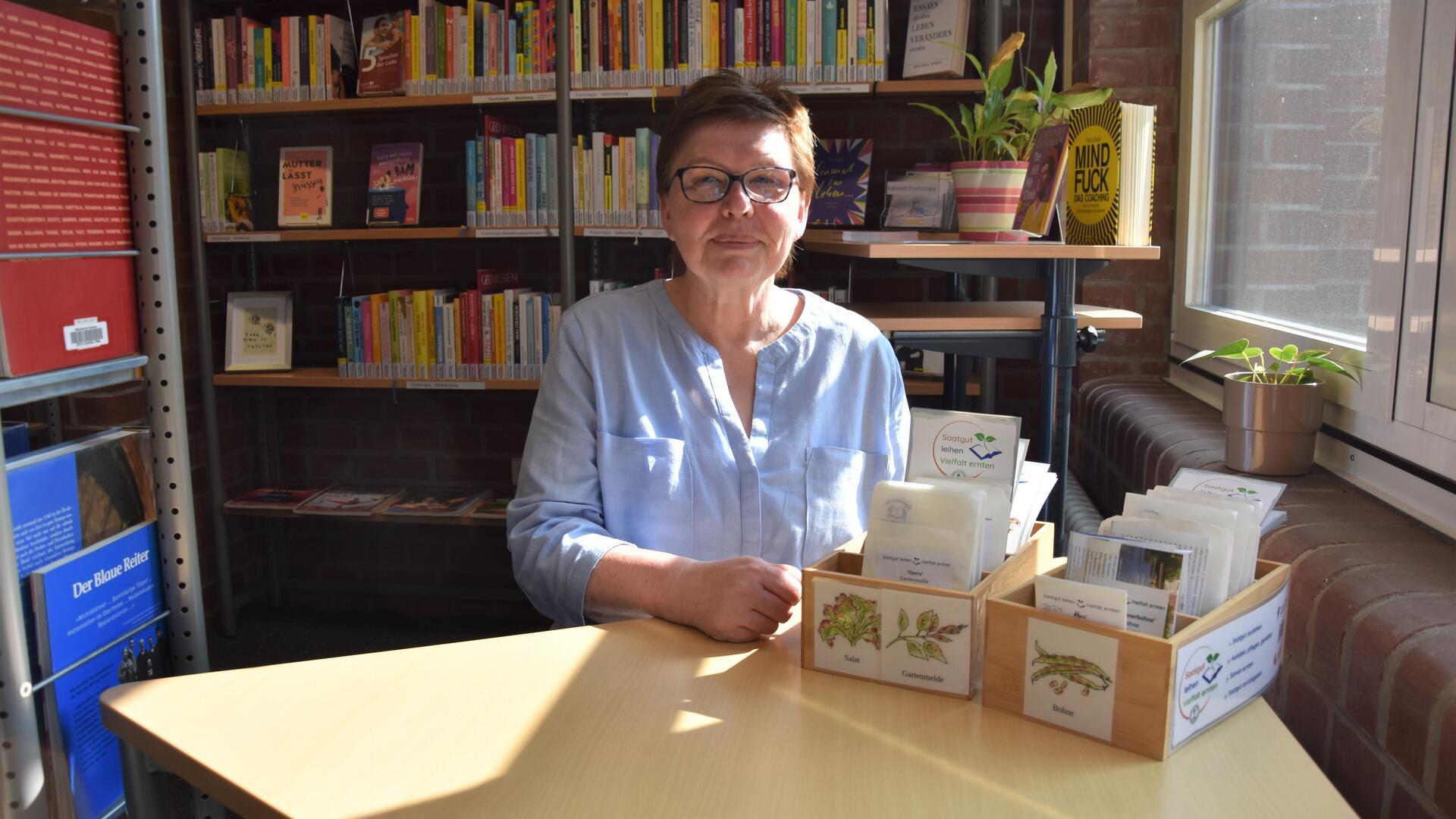 Eine Frau mit Brille sitzt vor Bücherregalen, und auf einem Tisch vor ihr stehen hölzerne Kisten mit kleinen Papiertüten.